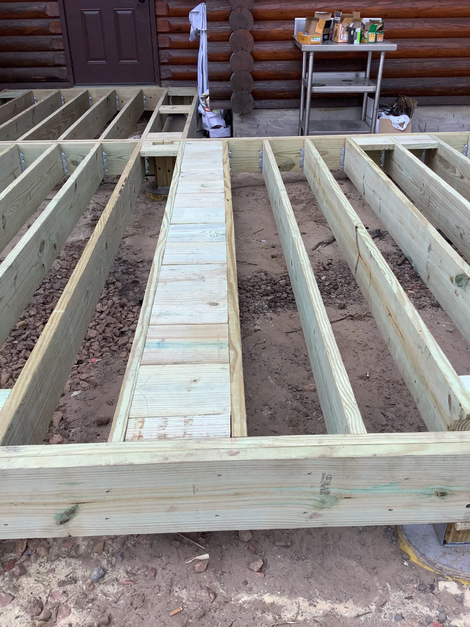 Wooden floor joists and beams laid out for deck construction next to a log cabin exterior door.