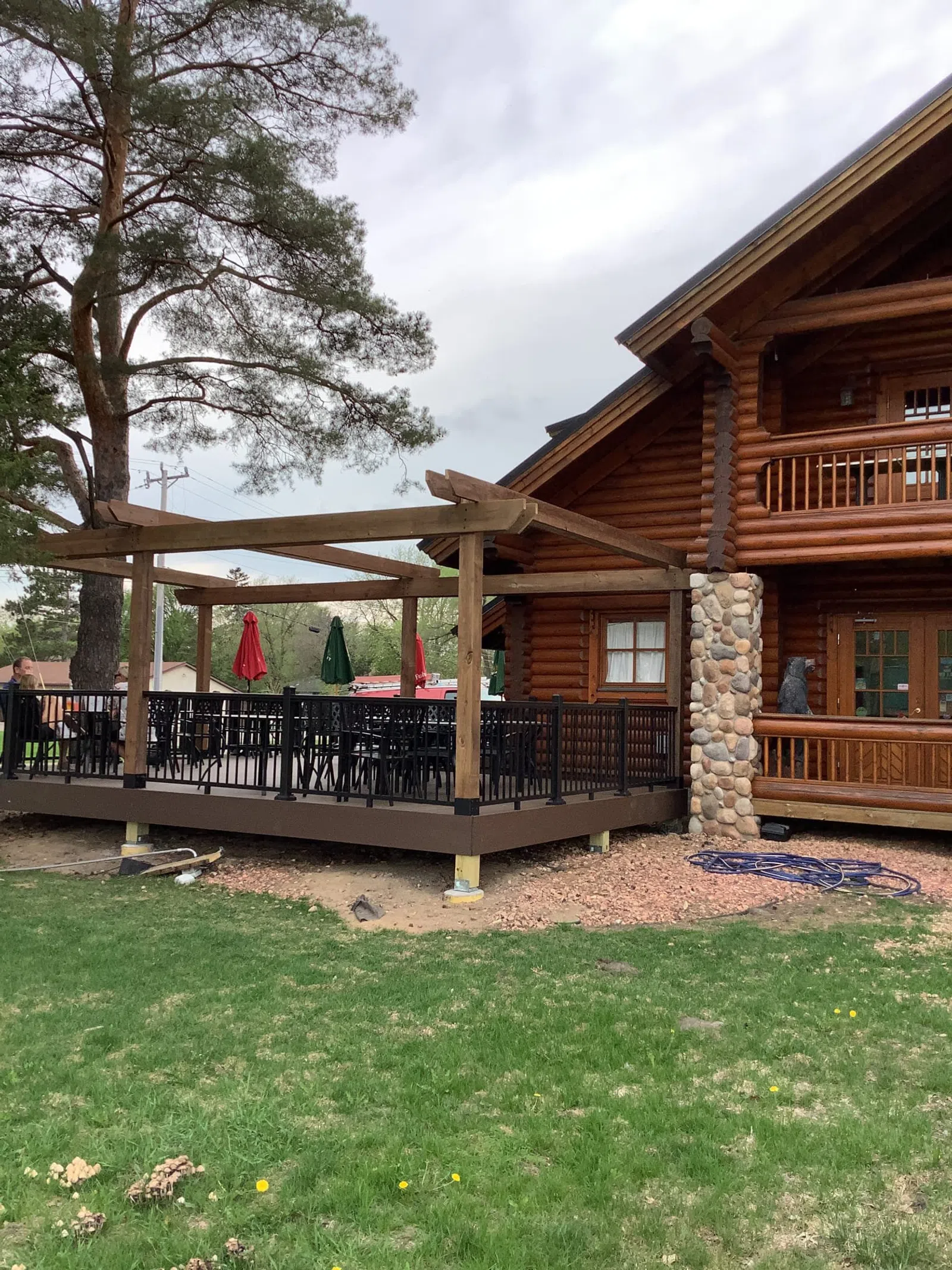 Outdoor wooden deck with black railing and pergola attached to a rustic log cabin surrounded by green grass.