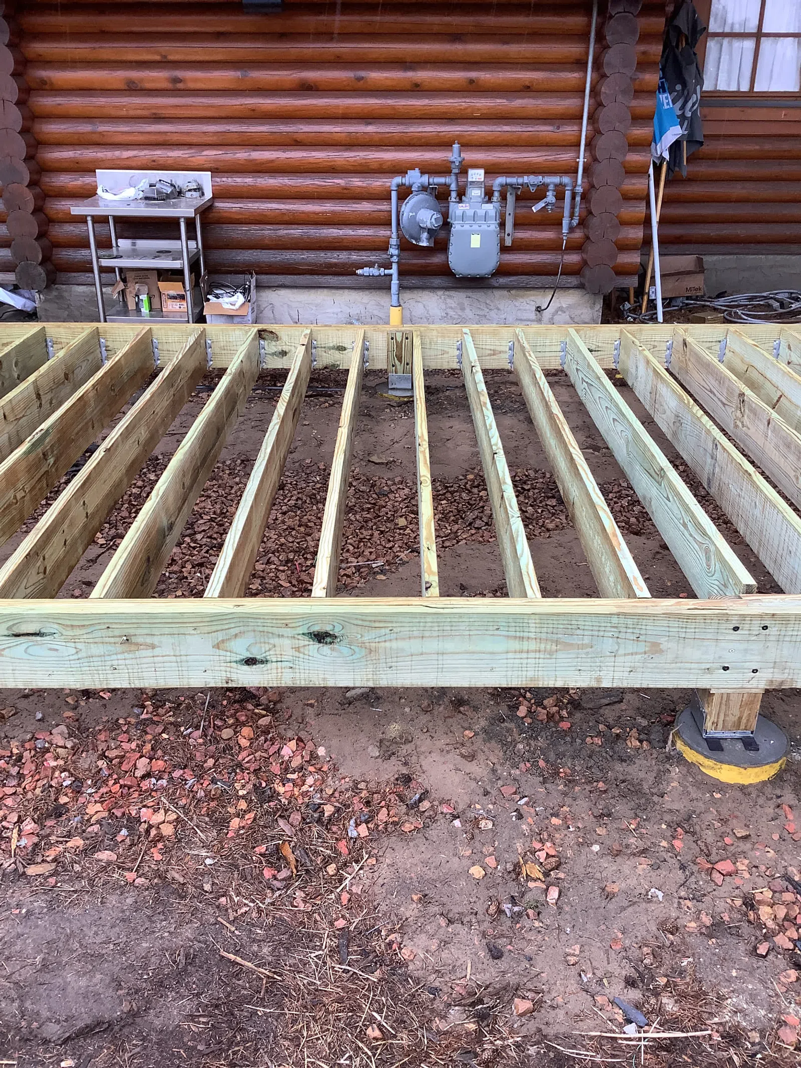 Wooden deck frame under construction in front of a log cabin with soil and gravel ground.