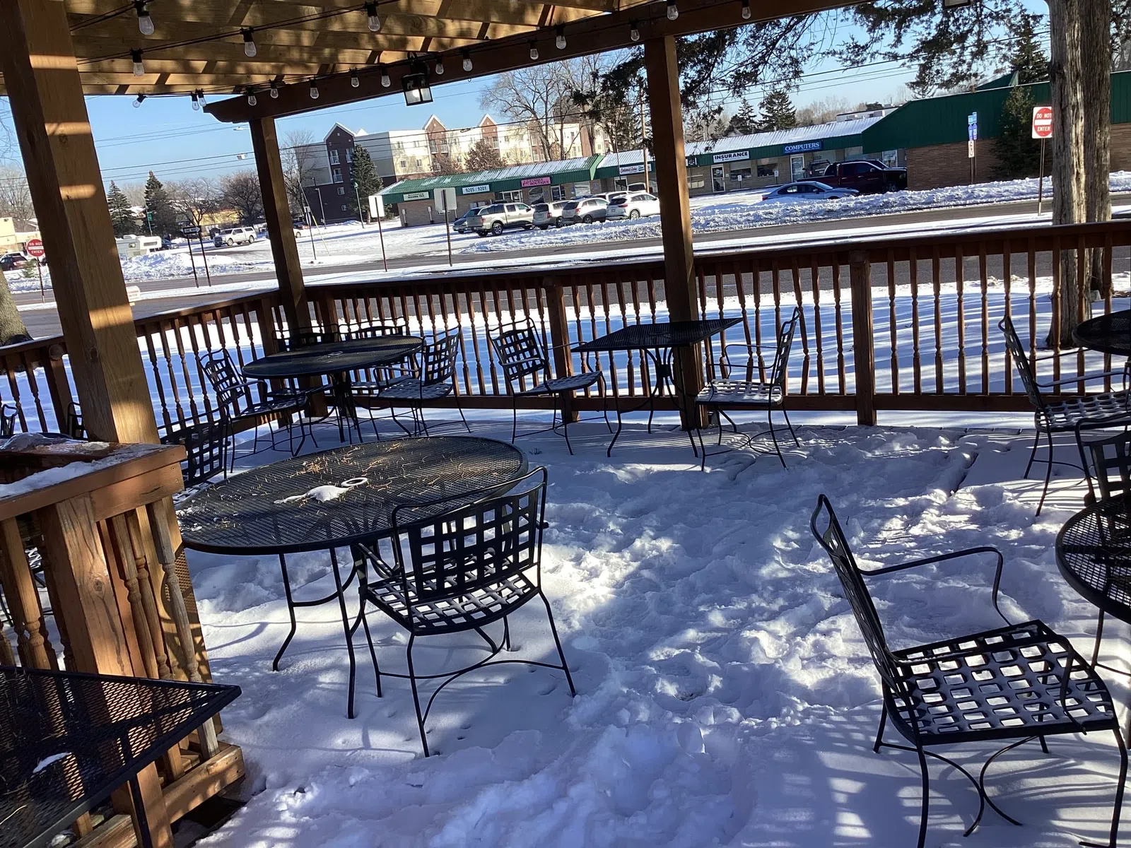Snow-covered outdoor patio seating with metal tables and chairs under wooden pergola on a sunny winter day.