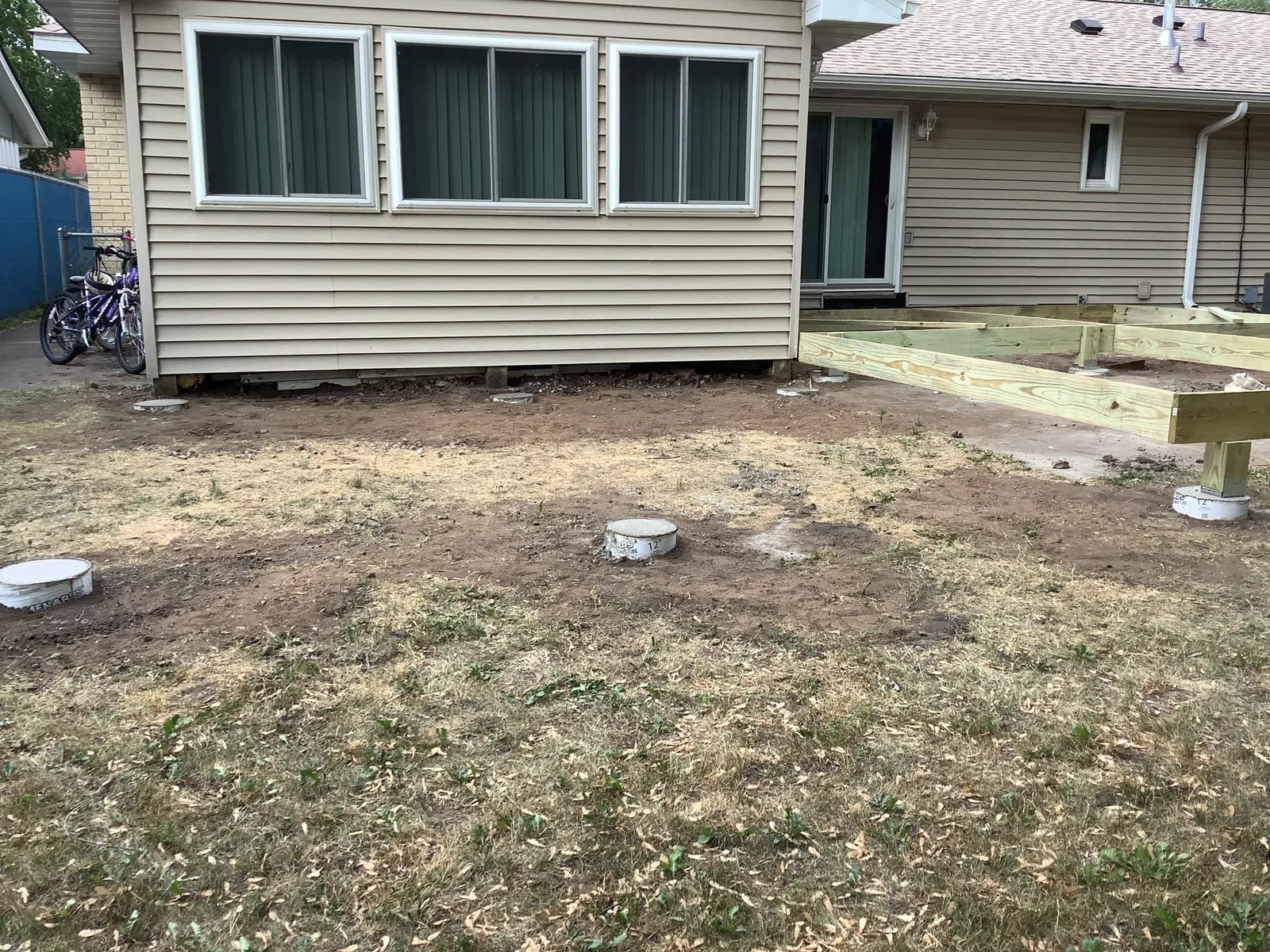 Backyard showing concrete pier foundations and wooden framing for a deck construction beside a house.