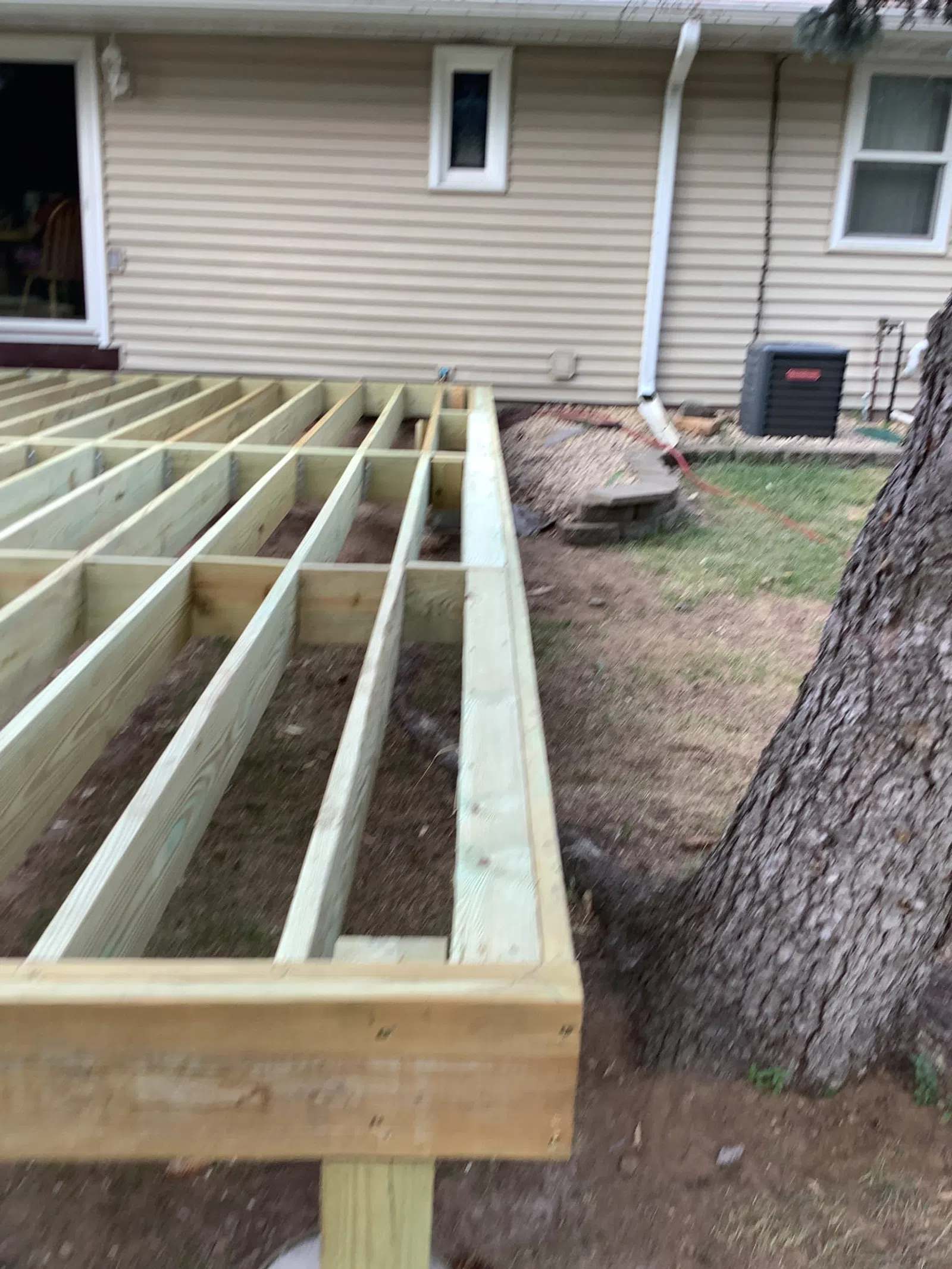 Partially constructed wooden deck frame next to a tree outside a house with beige siding.