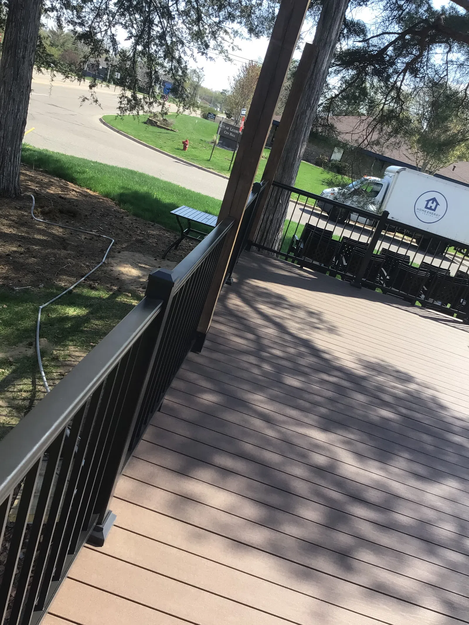 Spacious backyard deck with brown wooden flooring, black metal railing, and a white truck parked nearby on a sunny day.