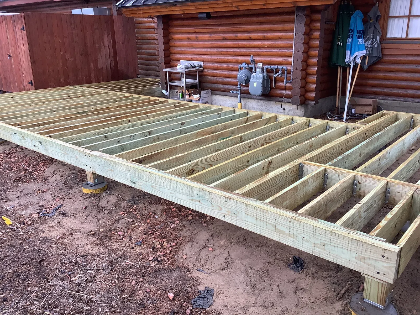 Wooden deck frame construction beside a log cabin on dirt ground, with tools and gas meters nearby.