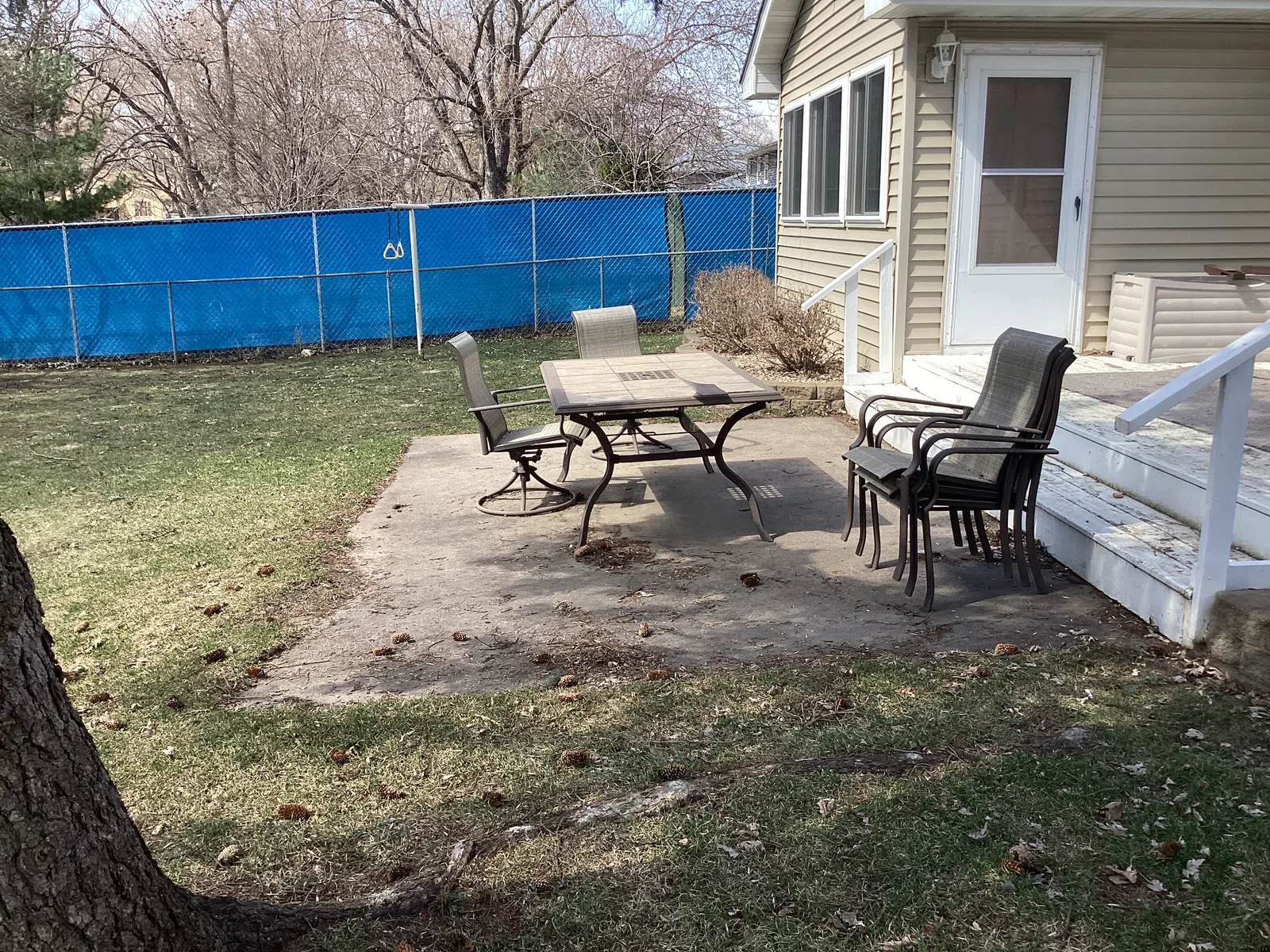 Backyard patio with outdoor table, four chairs, and house stairs under clear sky, fenced by blue mesh screen.