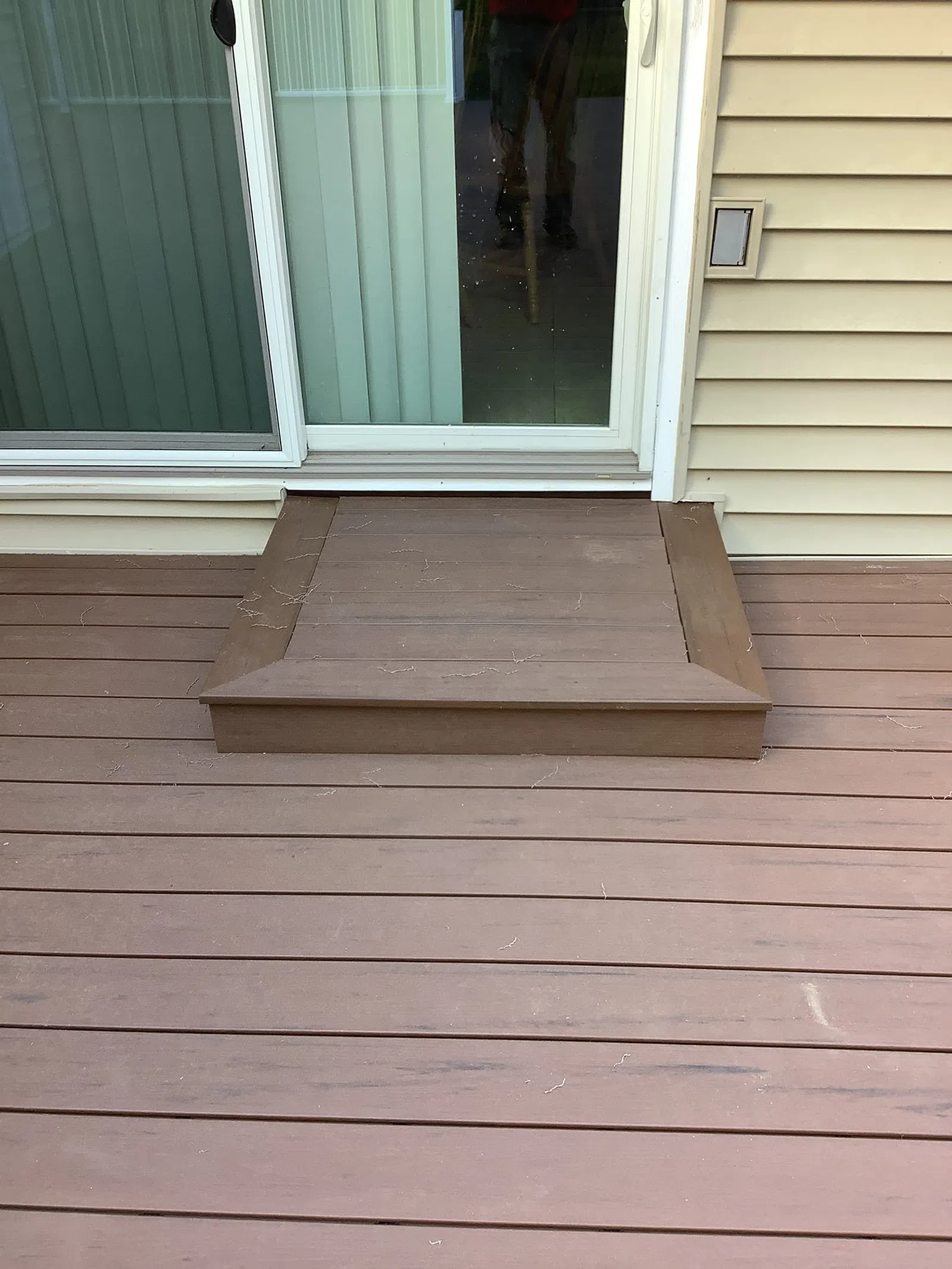 Brown wooden deck with a single step leading up to sliding glass door and light beige siding on house.