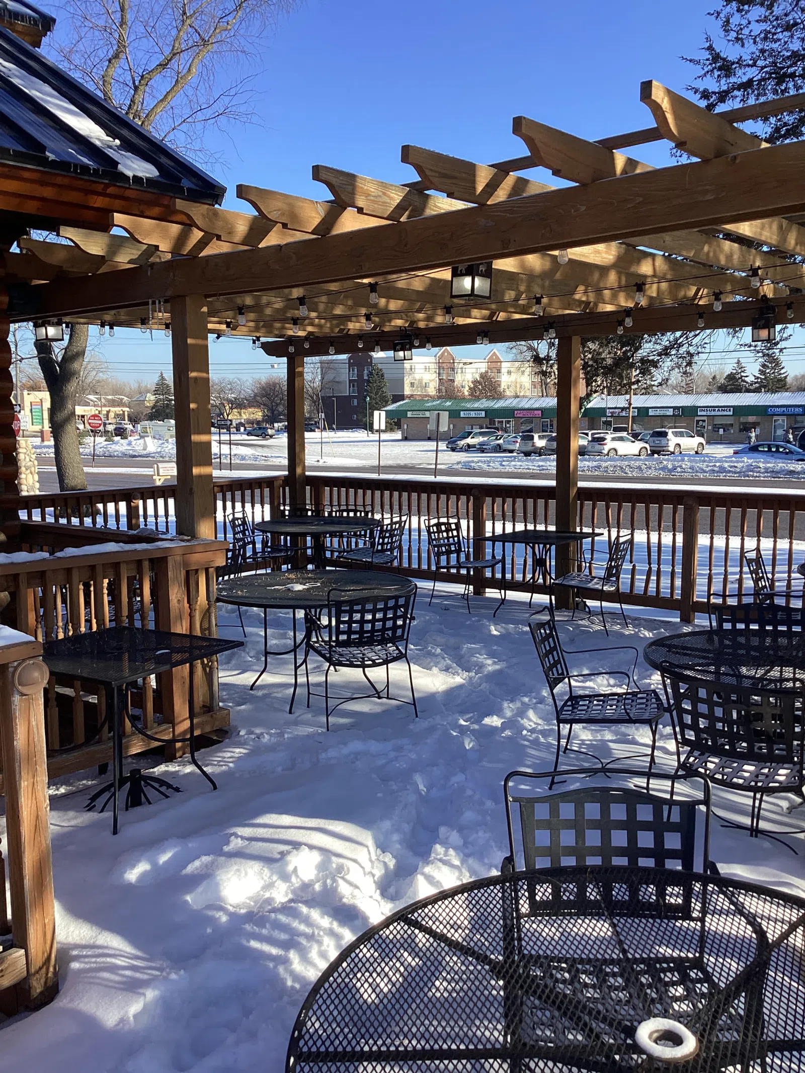 Outdoor patio with metal tables and chairs covered in snow under a wooden pergola on a bright winter day.