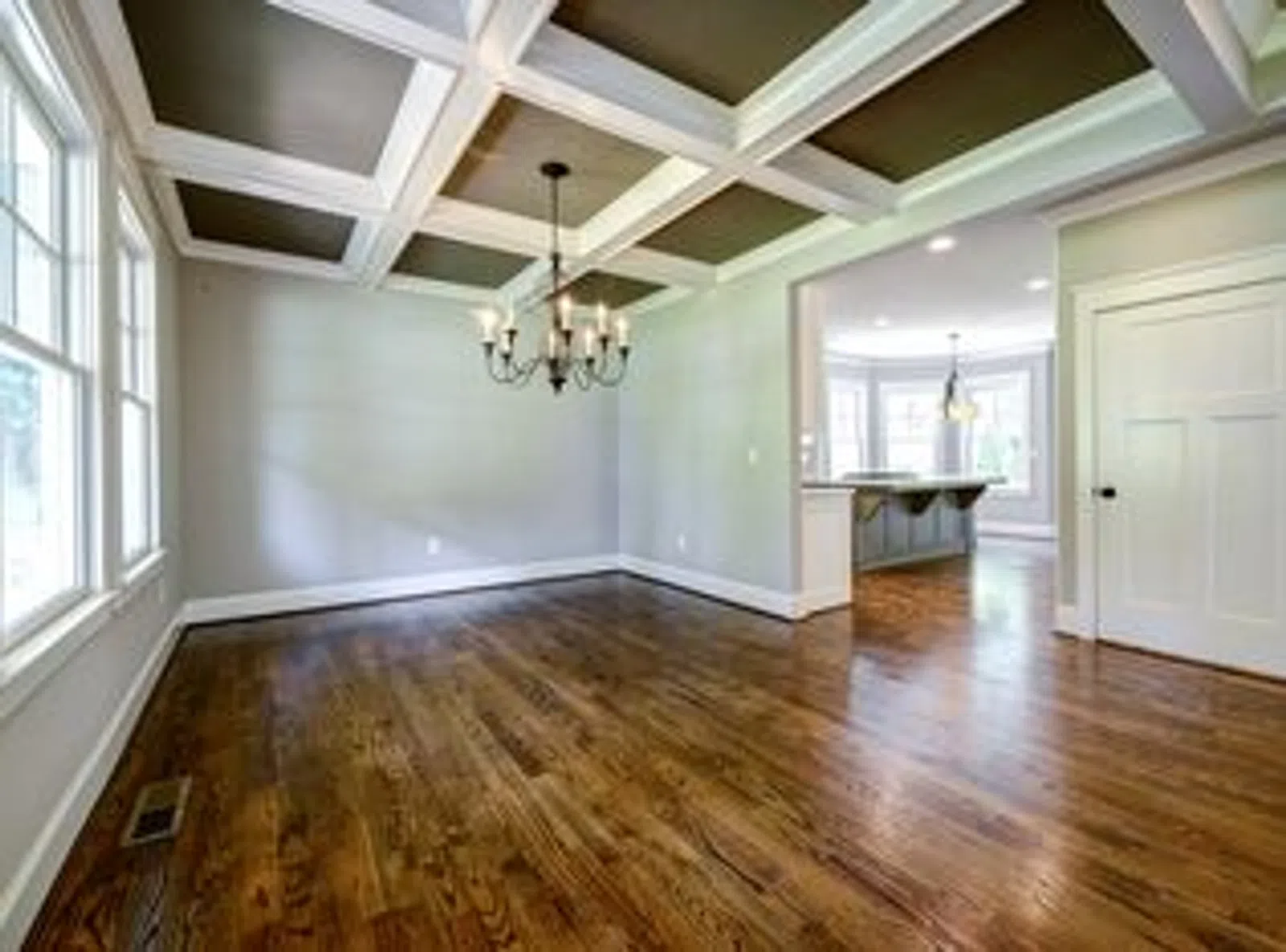 Empty room with hardwood floors, large windows, coffered ceiling, and chandelier lighting in a modern house.