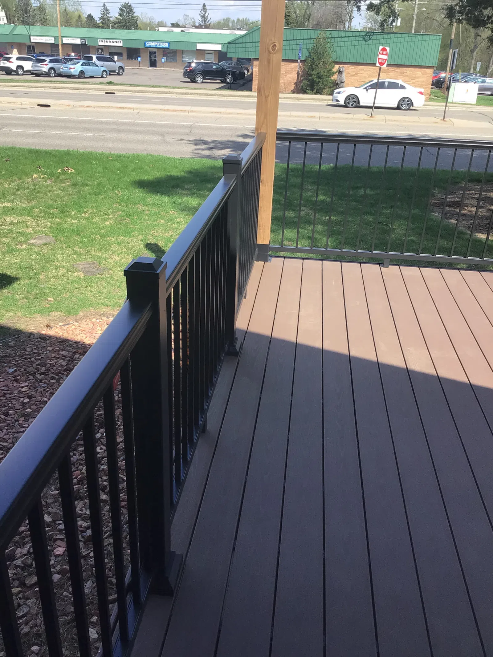 Outdoor deck with dark brown wooden floorboards and black metal railing overlooking a road and green lawn.
