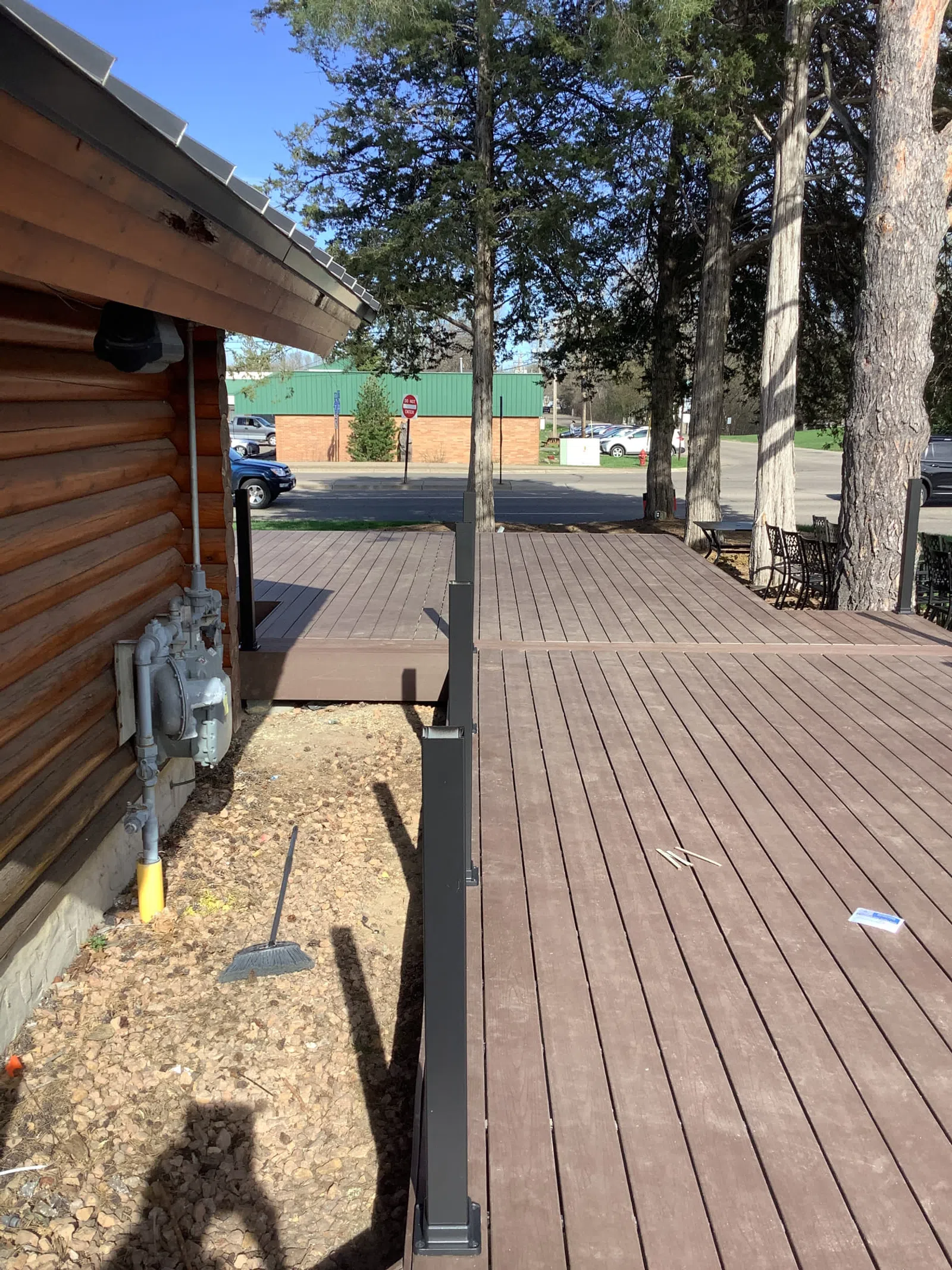 Wooden deck adjacent to a log cabin with metal railing, trees, broom, and parked cars in the background.