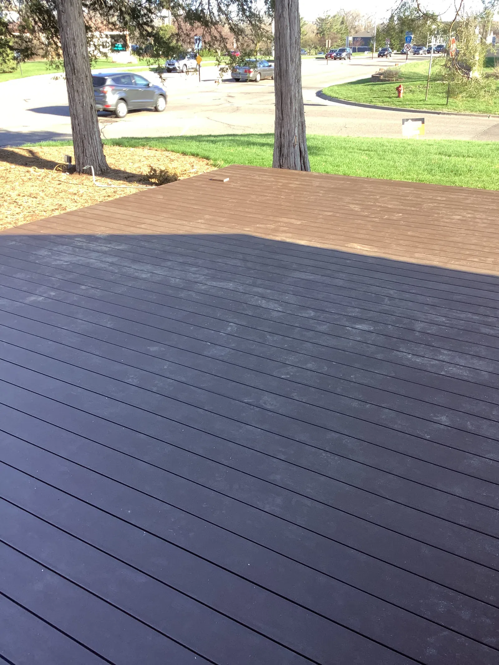 Two-tone wooden deck patio with black and brown planks under daylight near road and trees.