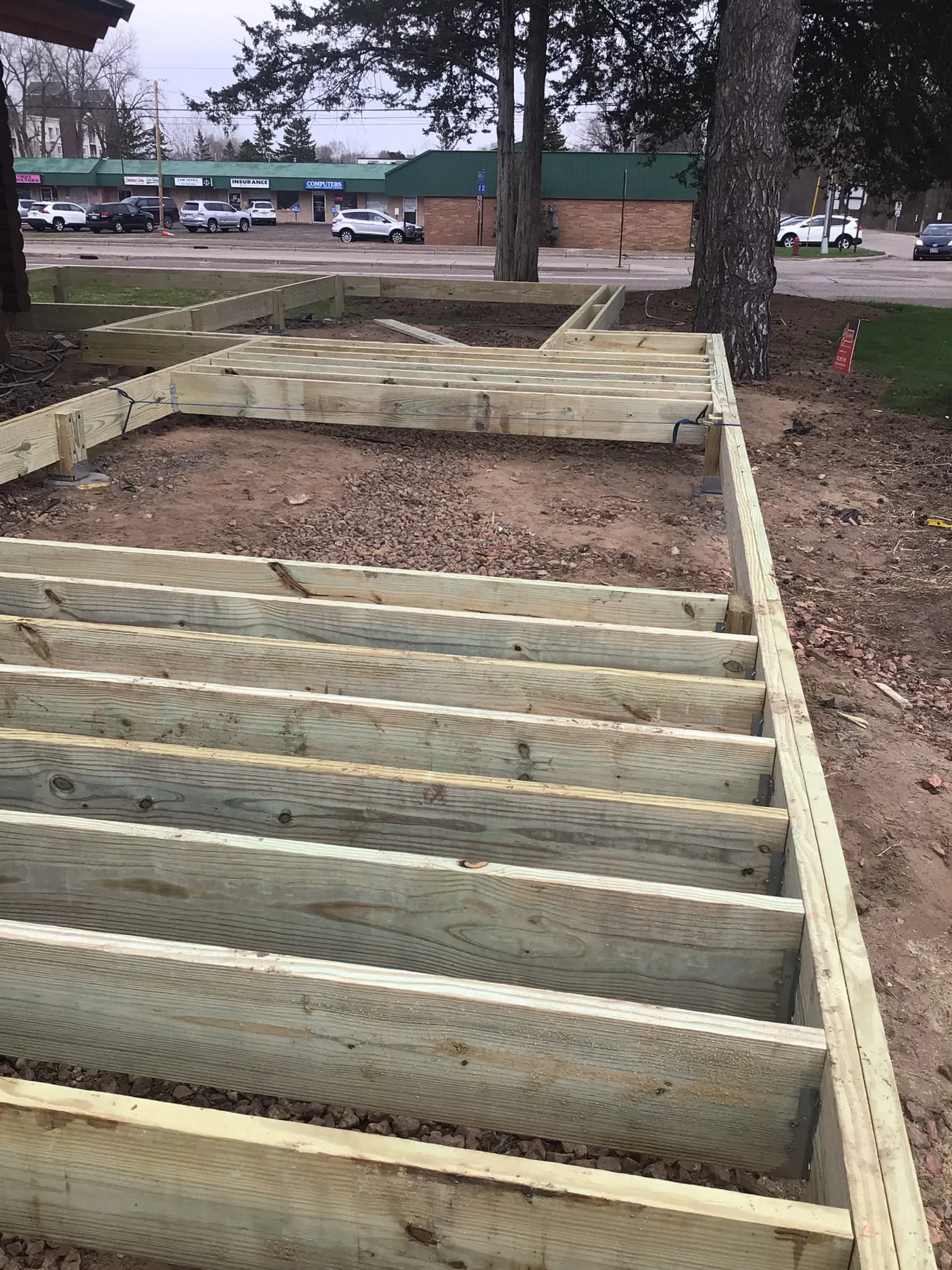Wooden deck frame under construction on dirt ground with nearby trees and road in the background.