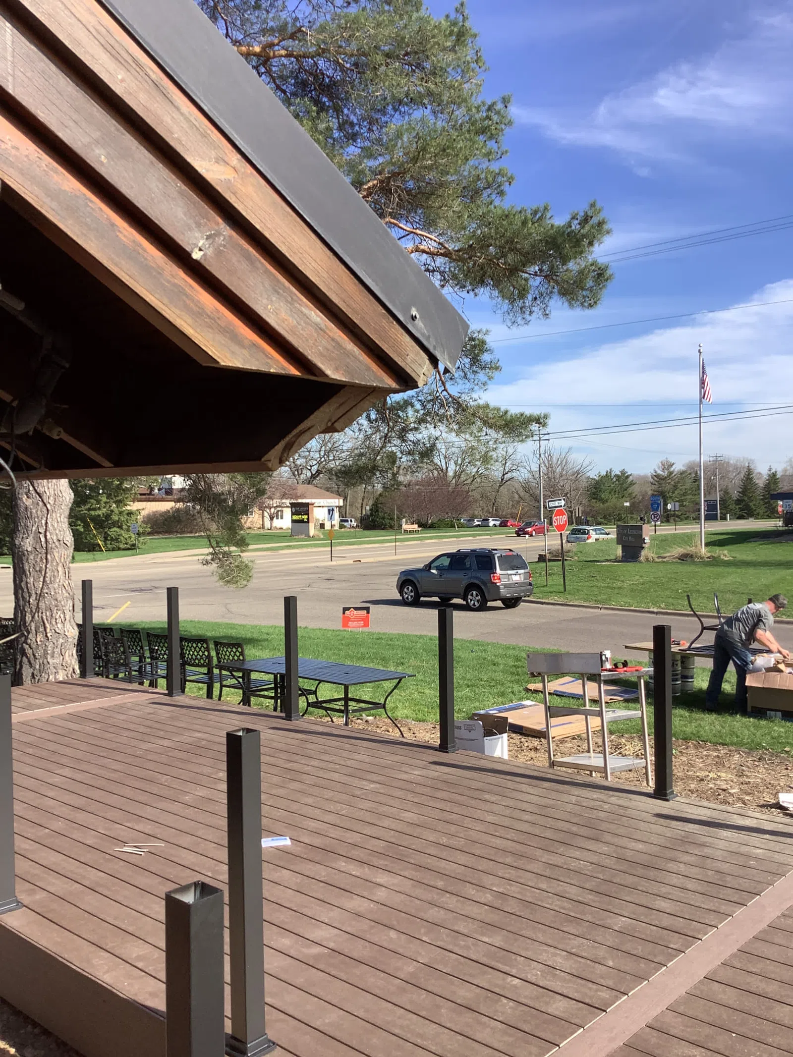Outdoor deck under construction near a road with a man working and a parked SUV on a sunny day.