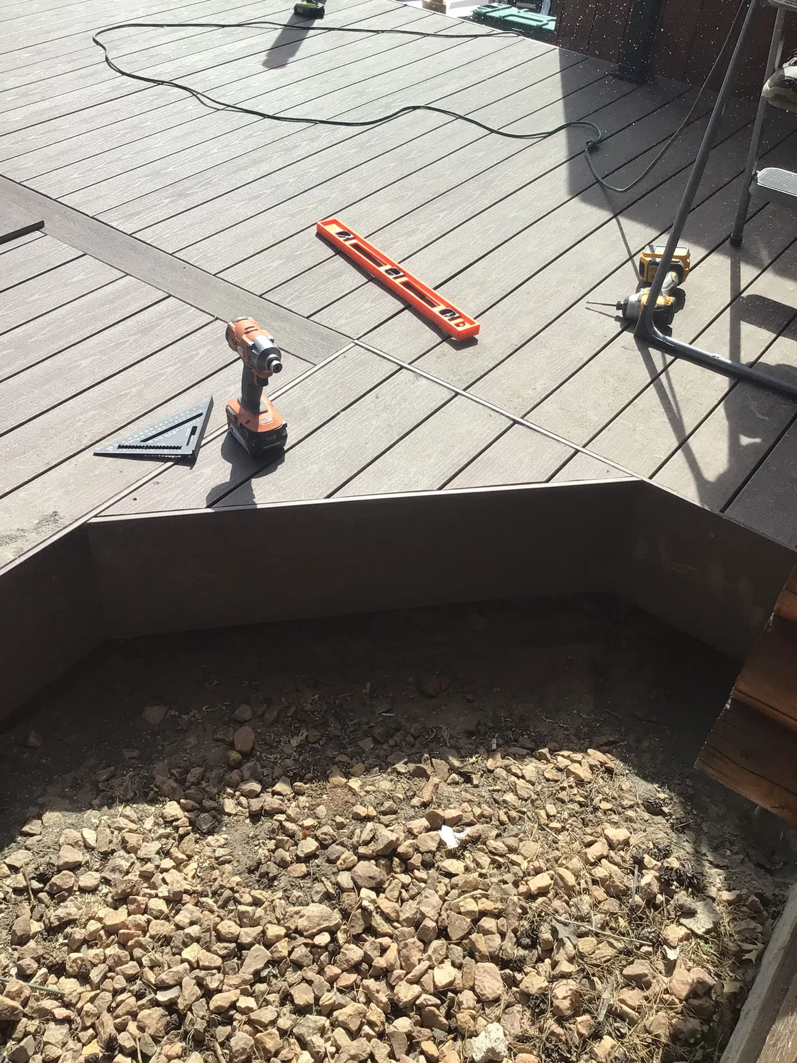 Wooden deck under construction with power drill, level, and rocky ground below the deck frame.