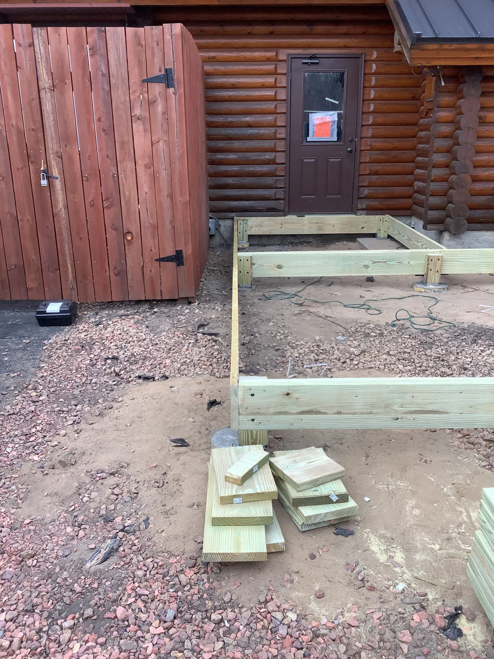 Construction of wooden deck frame beside log cabin with stacks of lumber and gravel ground