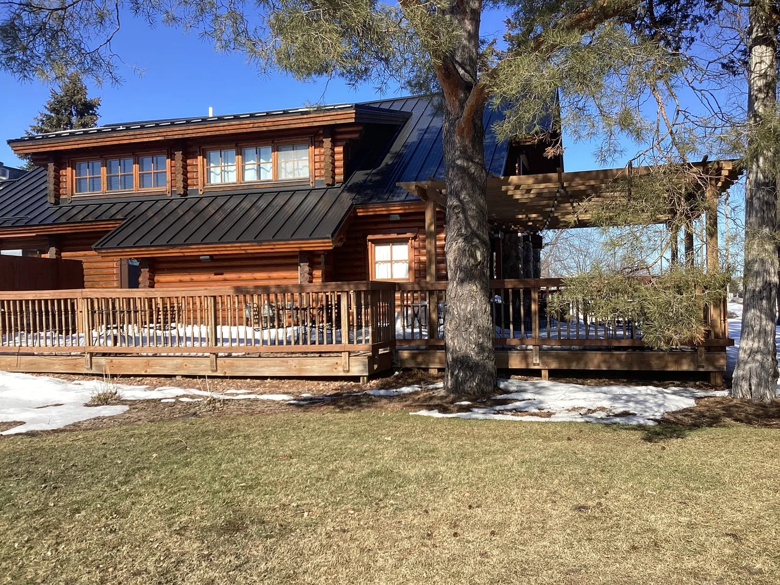 Wooden log cabin with black metal roof, large porch, trees, and patches of snow on the ground under clear blue sky