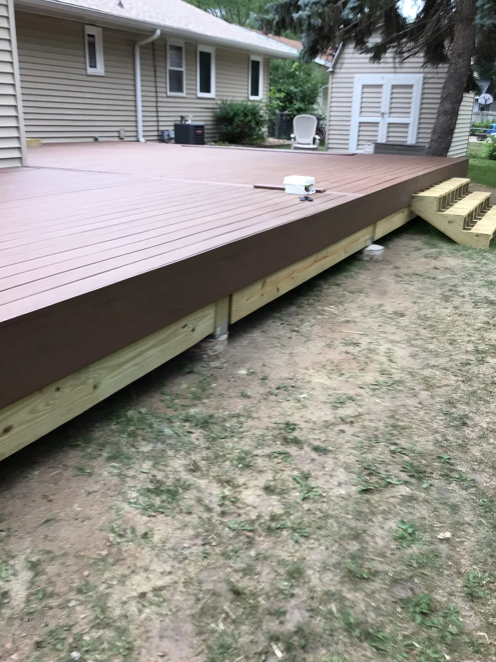 Newly built brown wooden backyard deck with stairs next to house and garden shed on grassy ground.