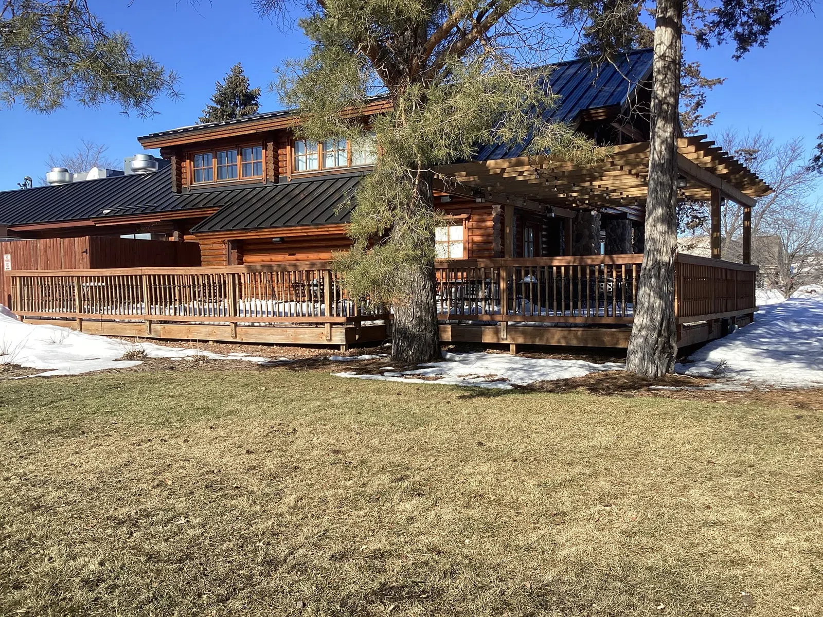 Rustic wooden cabin with black metal roof, large deck, surrounded by snow patches and trees on sunny day.