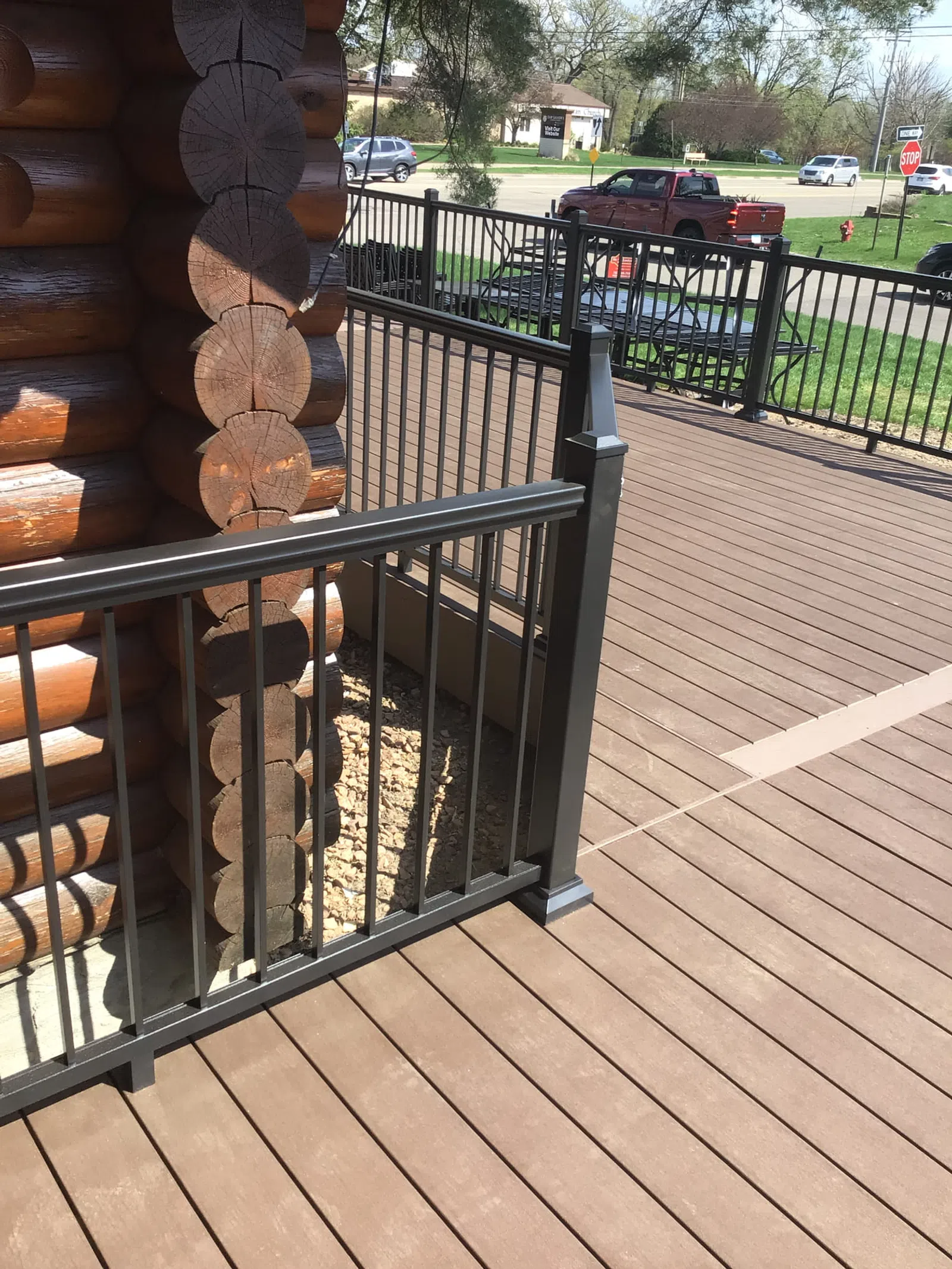 Brown wooden deck with black metal railing attached to a log cabin corner, outdoor suburban setting.