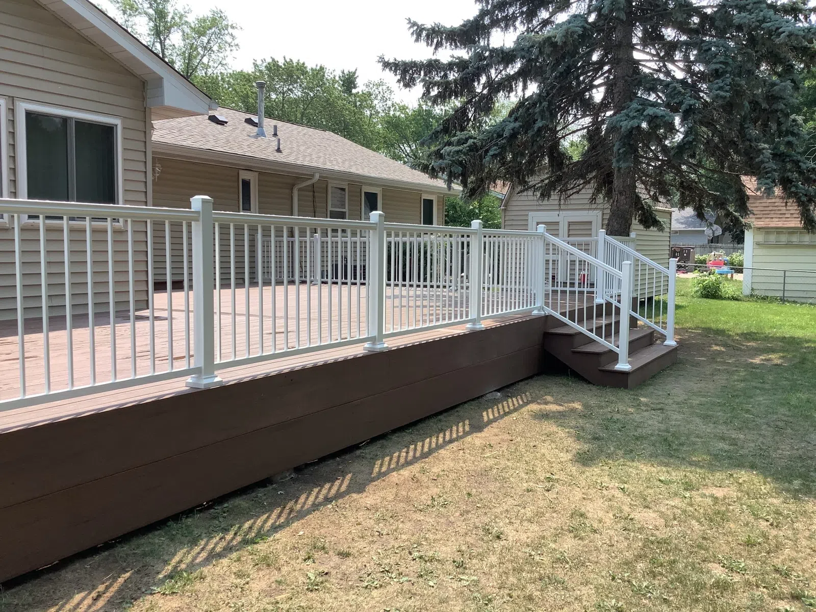 Wooden backyard deck with white railing and stairs attached to a beige house, surrounded by grass and trees.