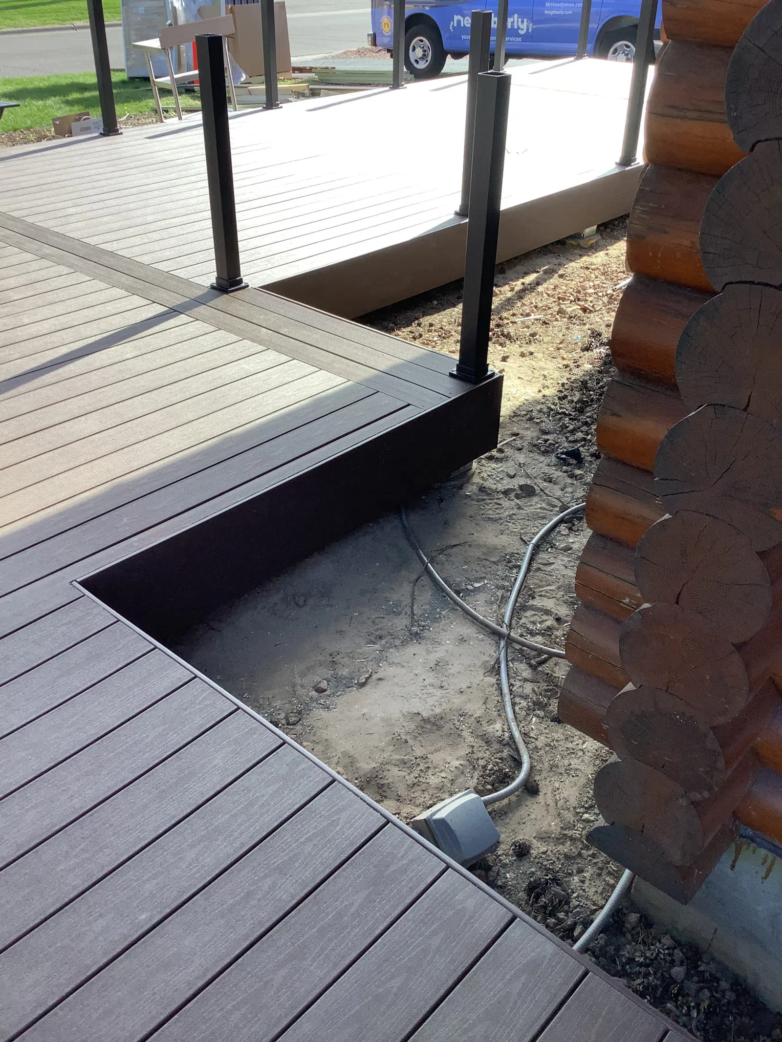 Close-up of brown composite decking and railing next to a log cabin corner with exposed ground and cables.