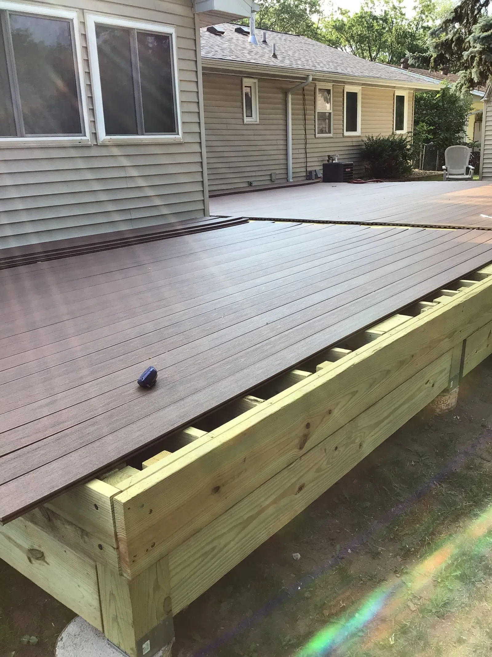 Partially installed dark composite decking boards on a wooden deck frame next to a house exterior in daylight.