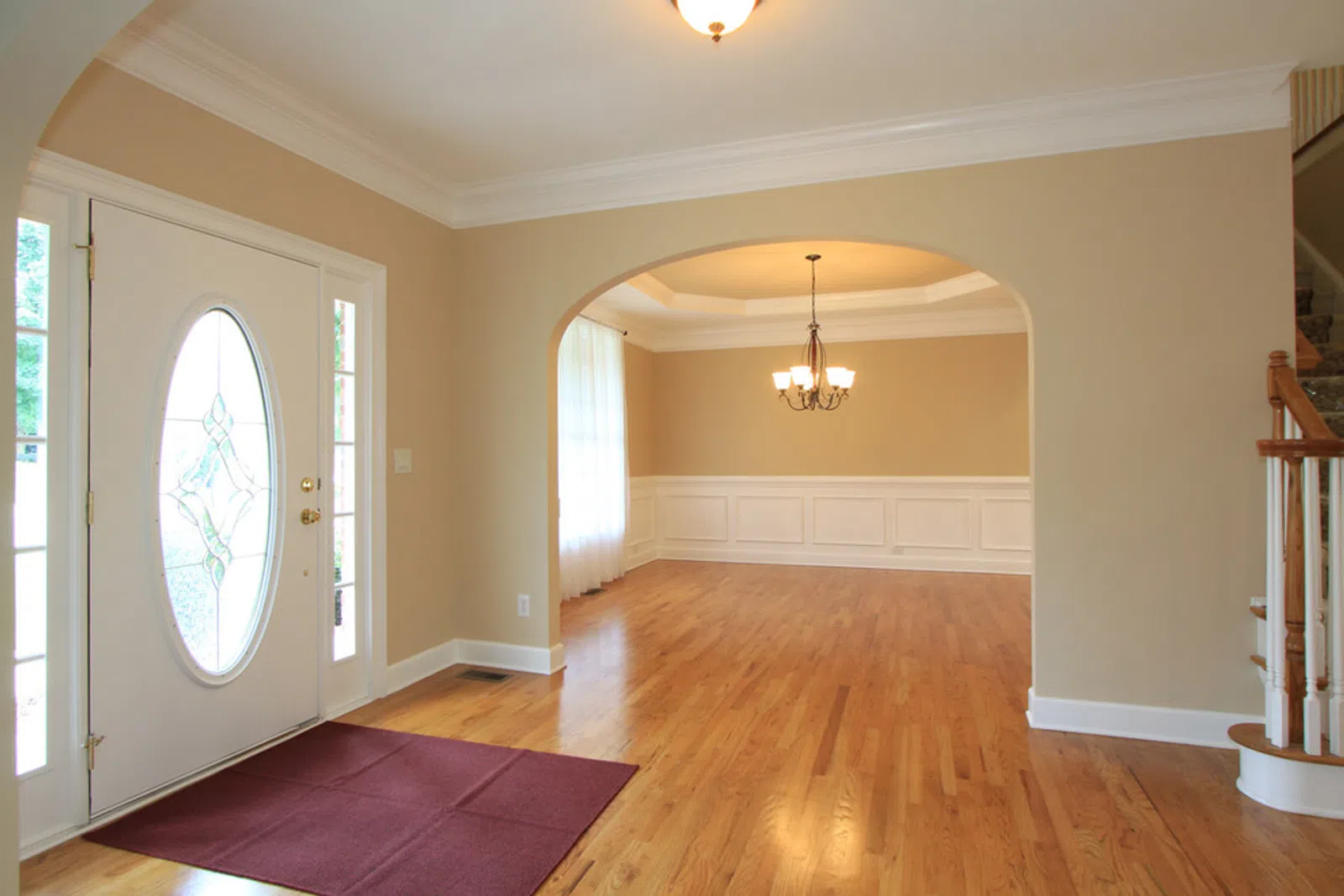 Bright empty living room with hardwood floors, beige walls, white trim, oval glass front door, and chandelier.