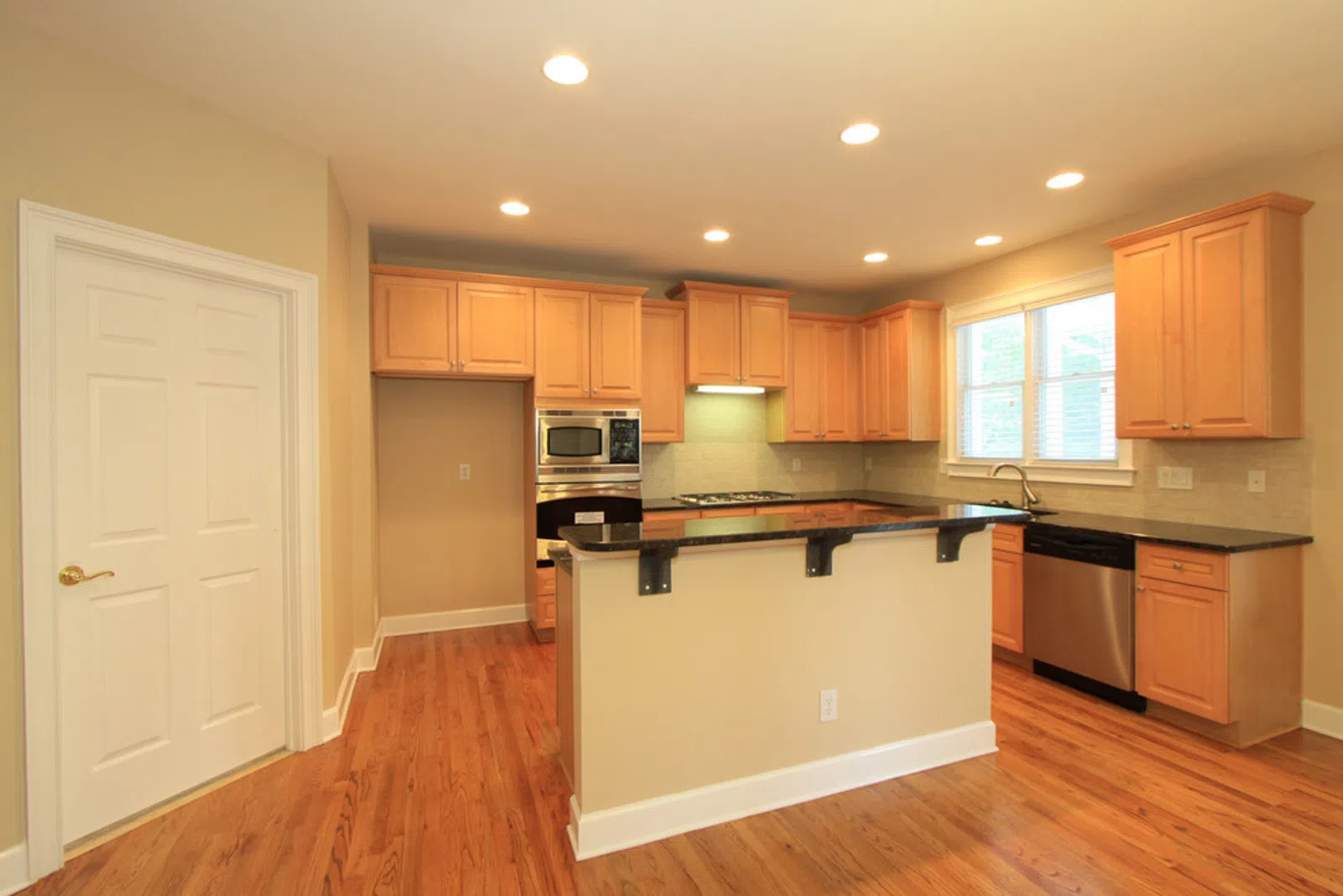 Bright modern kitchen with wooden cabinets, black countertops, stainless steel appliances, and hardwood floors.