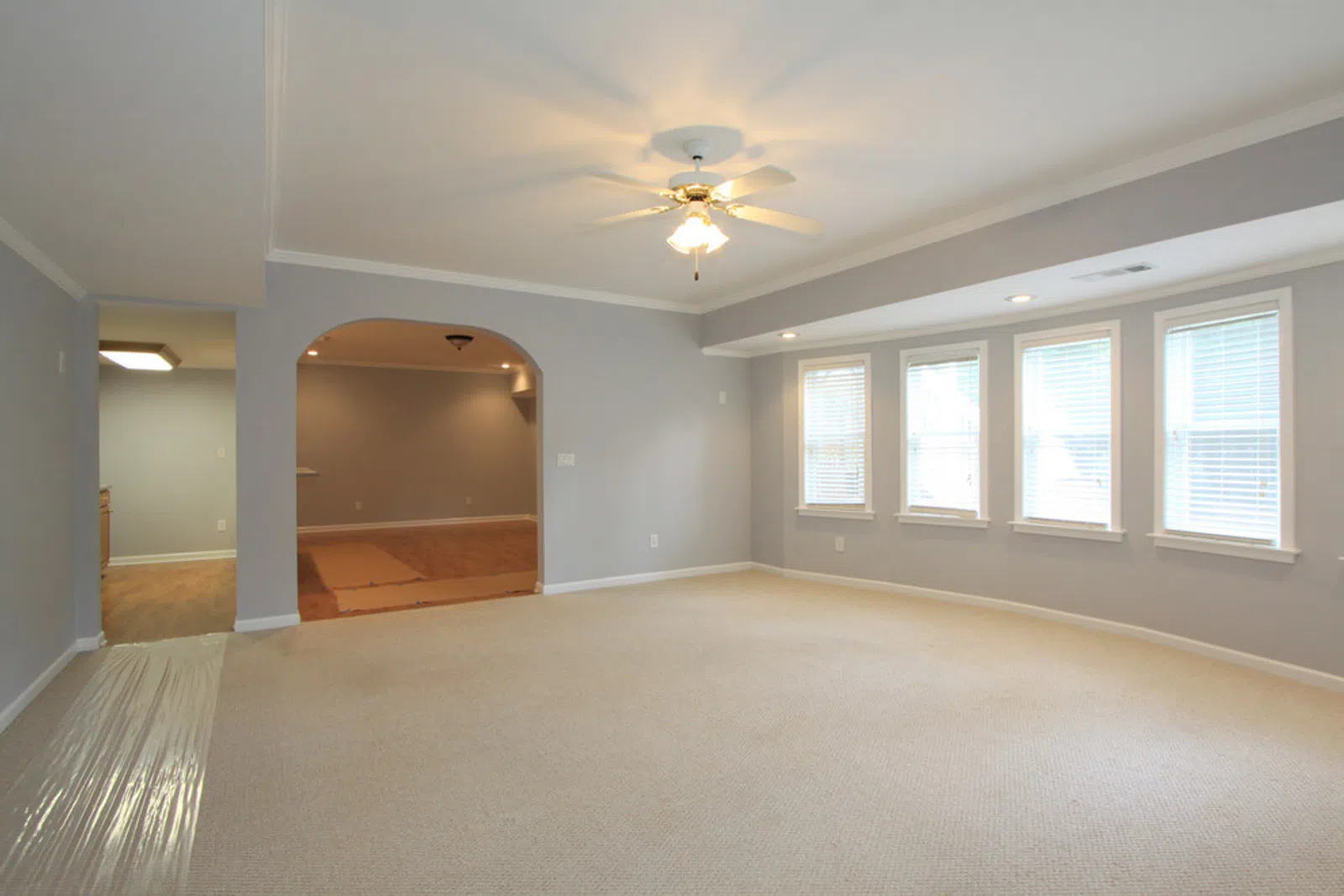 Bright empty living room with beige carpet, gray walls, ceiling fan, and three large windows with blinds.