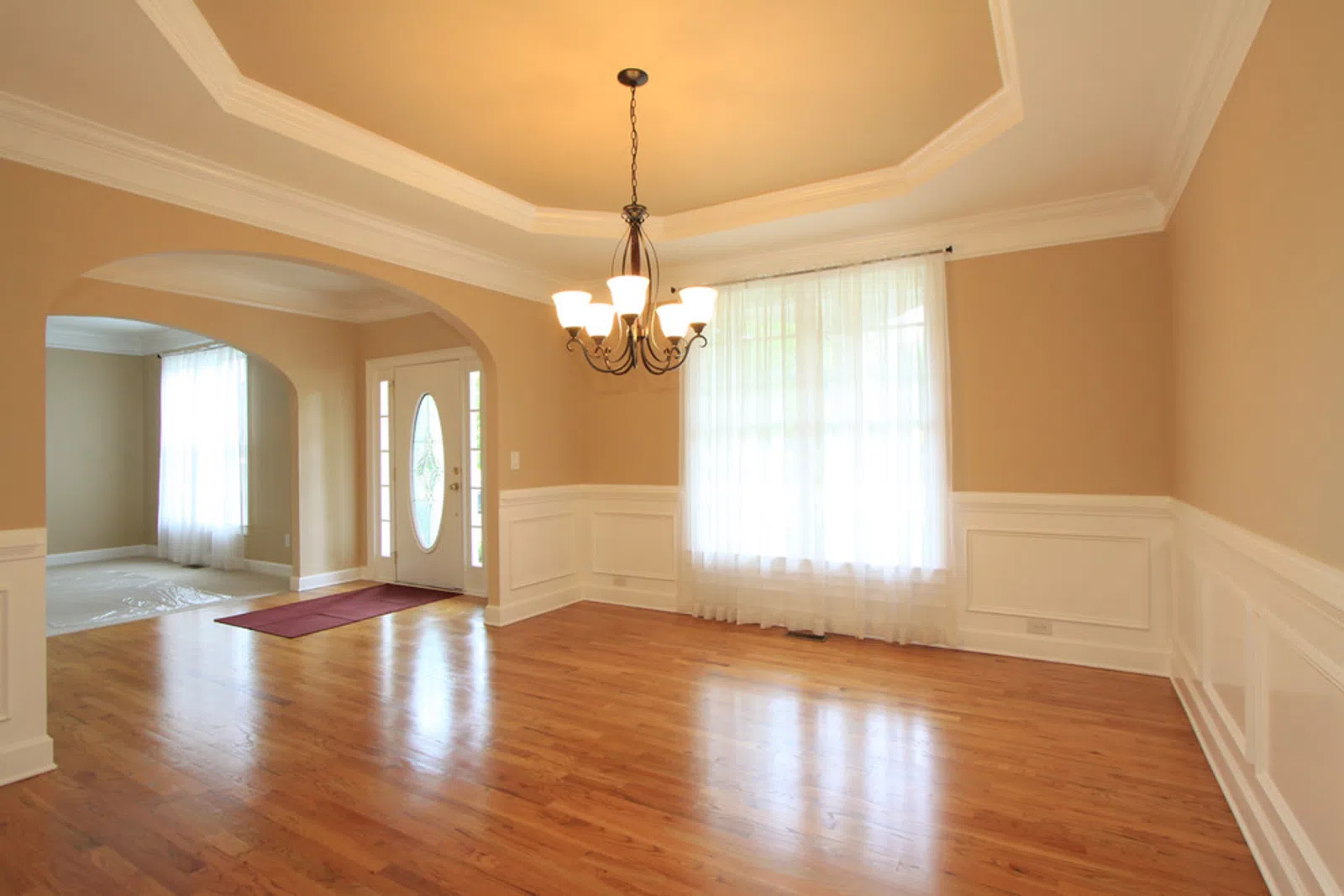 Empty dining room with hardwood floors, beige walls, white wainscoting, chandelier, and a window with sheer curtains