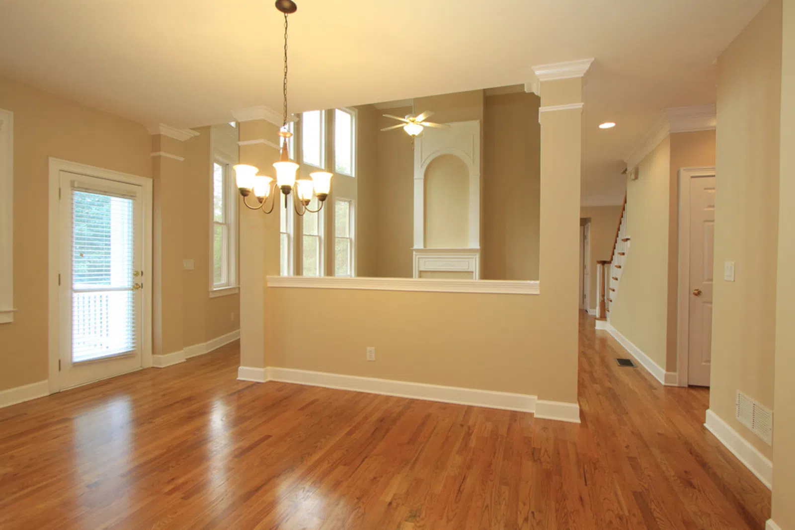 Empty spacious room with hardwood floors, beige walls, chandelier, ceiling fan, and large windows letting in natural light