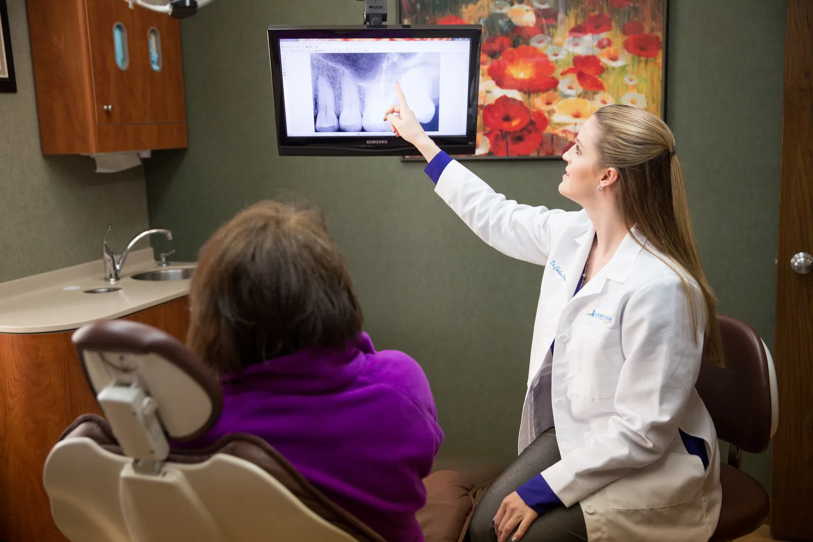 Dentist showing dental X-ray on screen to patient seated in dental office chair with colorful wall art.