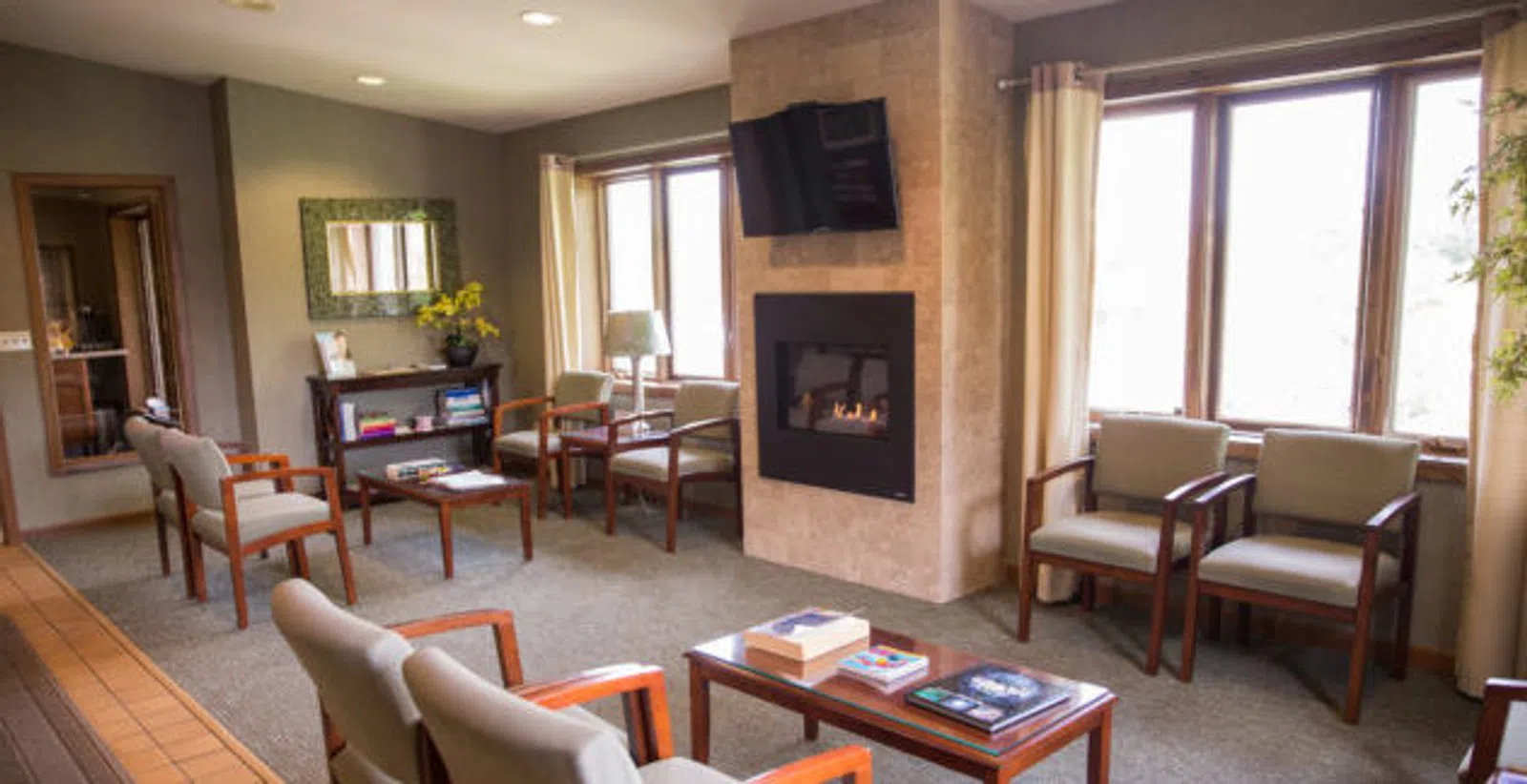 Cozy waiting room with cushioned chairs, a fireplace, flat-screen TV, and natural light from large windows.