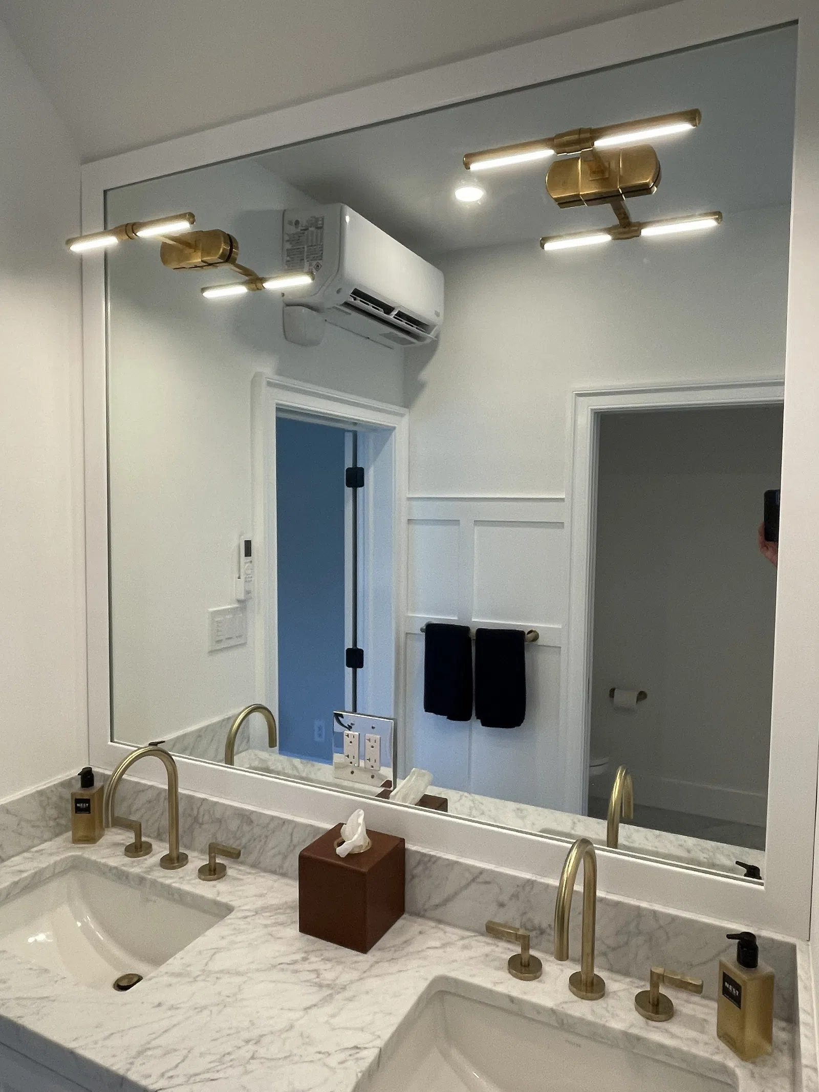 Modern bathroom with dual sinks, marble countertop, brass faucets, large mirror, and wall-mounted air conditioner.