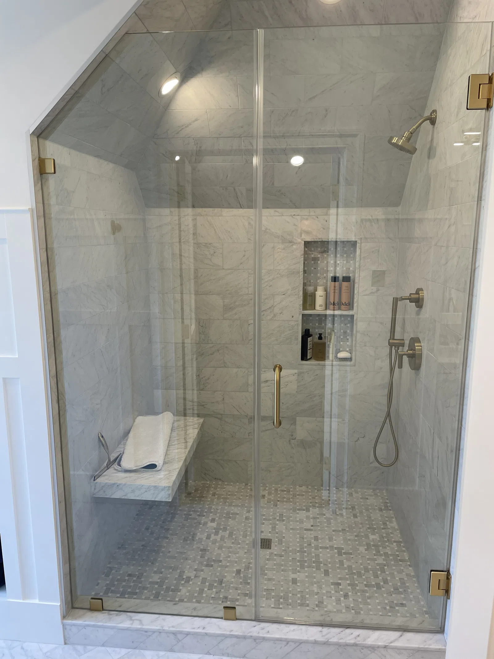 Modern marble tiled walk-in shower with glass doors, built-in bench, and gold-tone fixtures and showerhead.