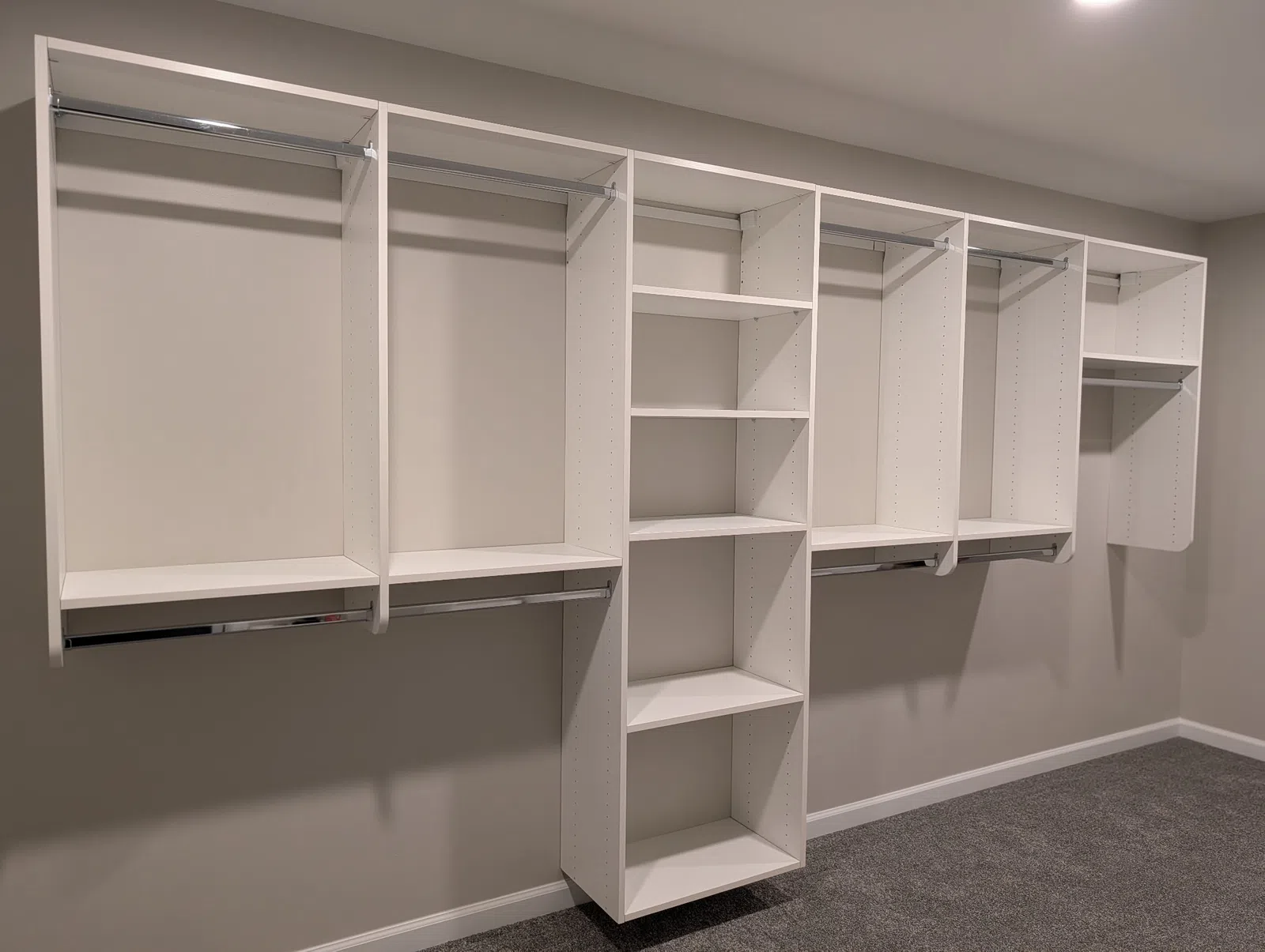 Empty white modular closet organizer with shelves and hanging rods against beige wall and carpeted floor.