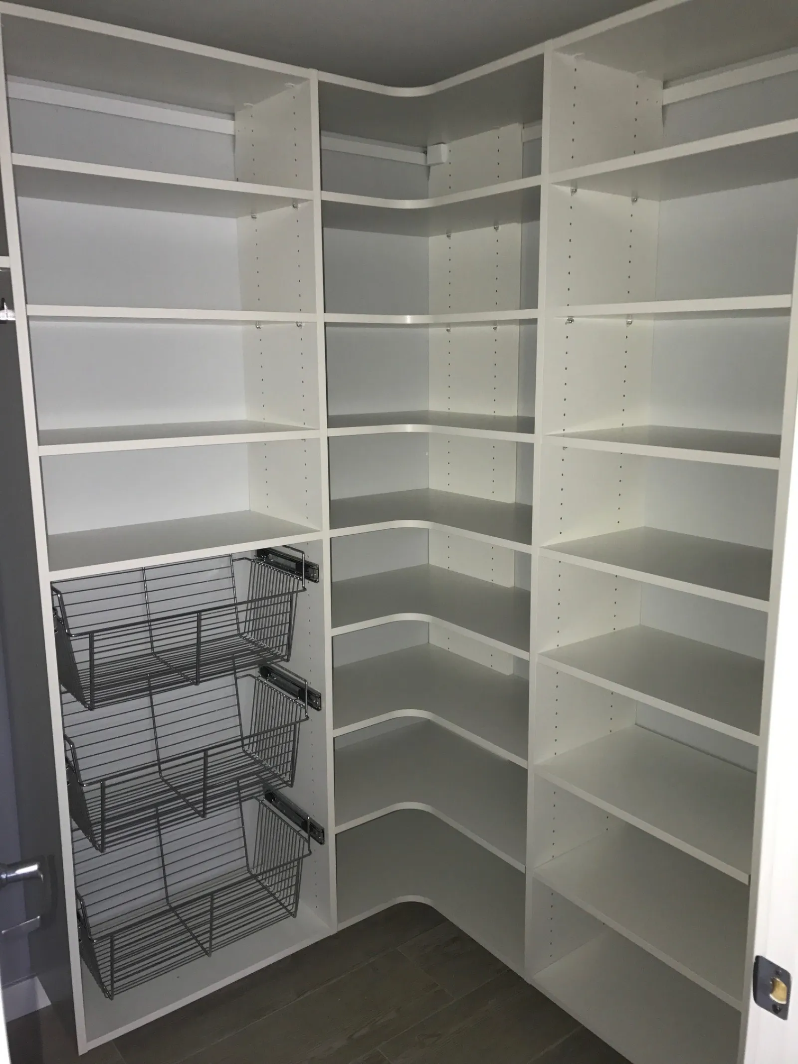 Empty white corner pantry with adjustable shelves and three metal wire baskets on the left side.