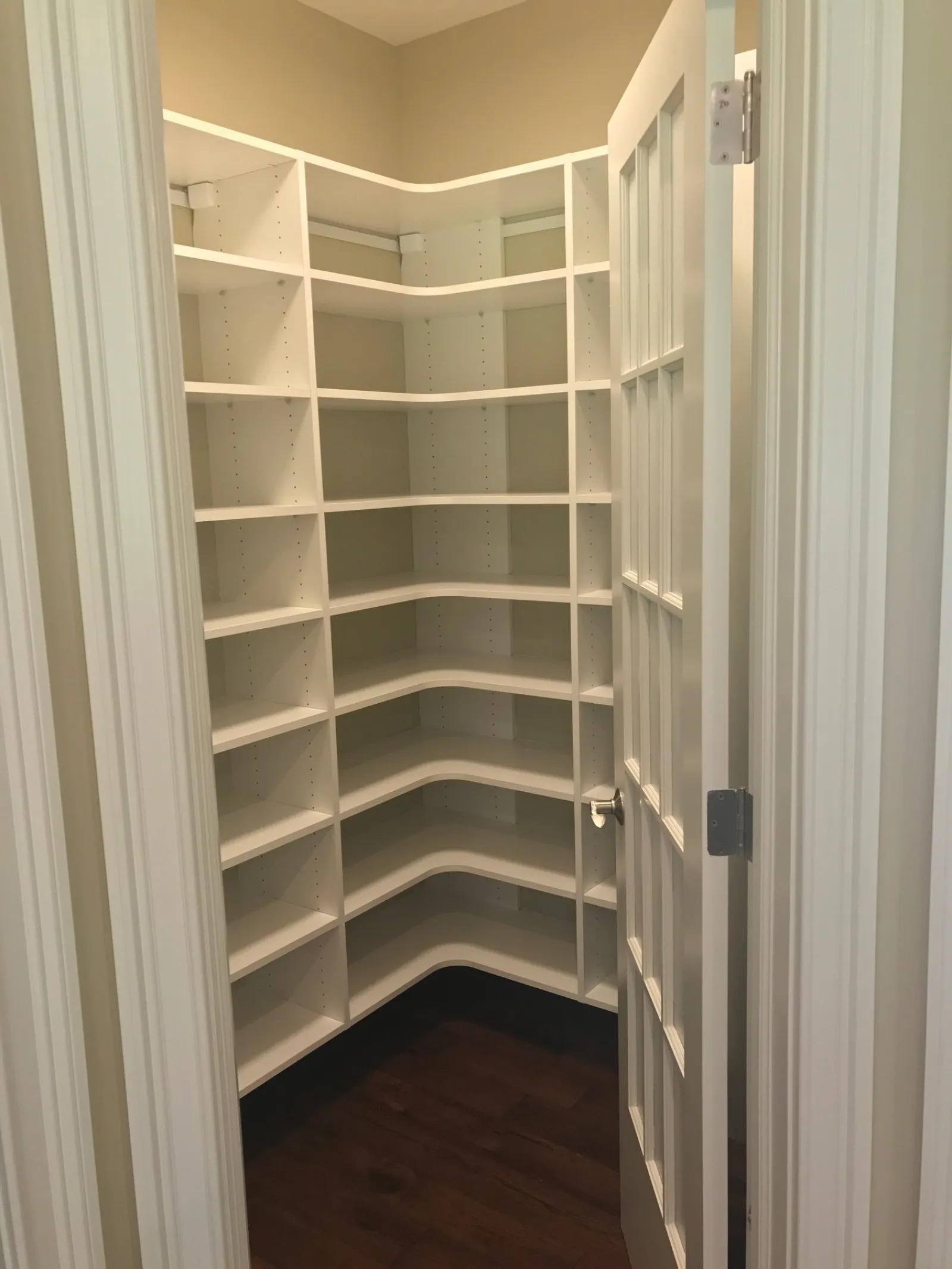 Empty white corner pantry shelves inside a small room with an open door and dark wooden floor