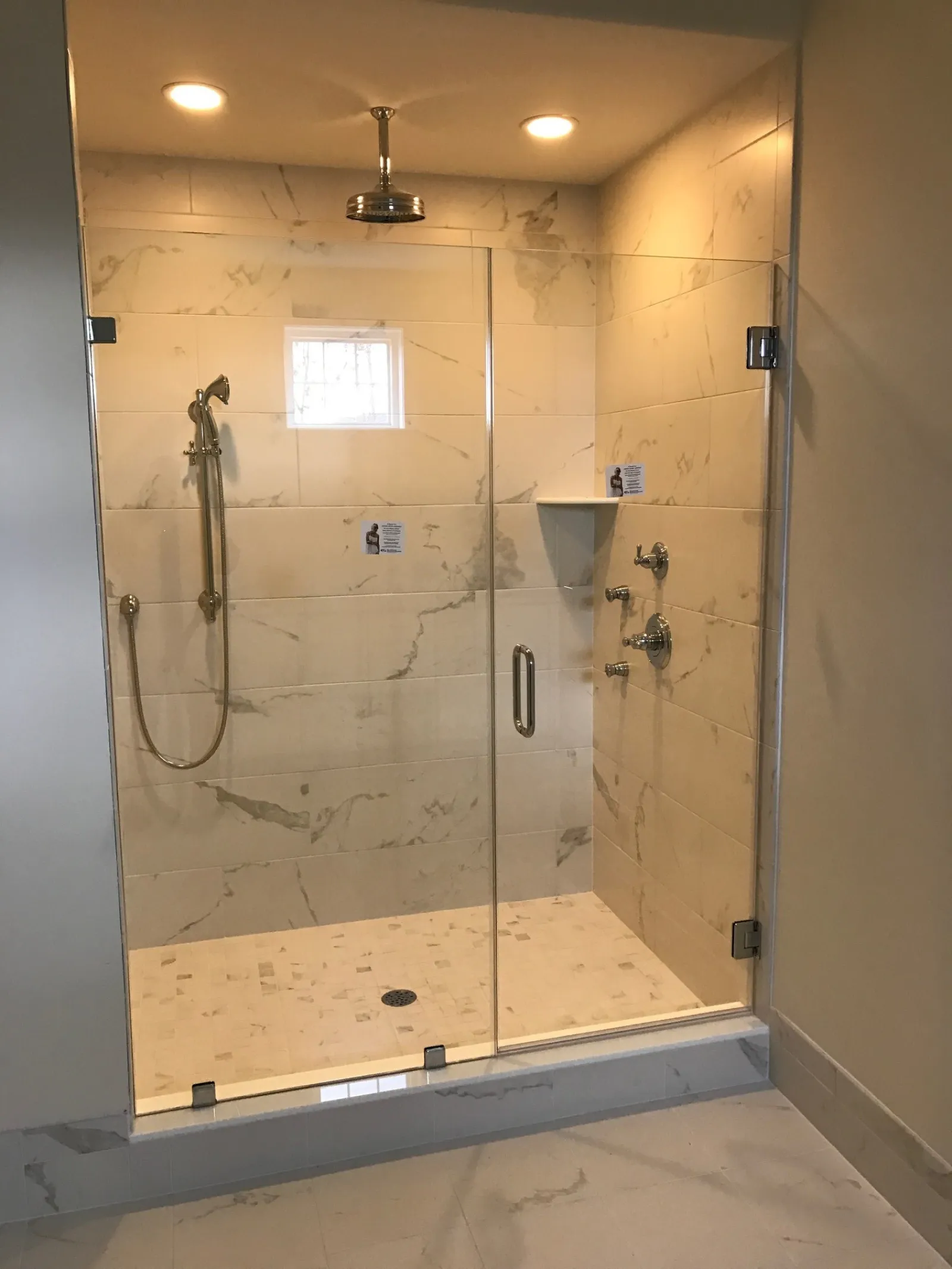 Modern walk-in shower with glass door, marble tiles, rain showerhead, and handheld shower in bathroom.