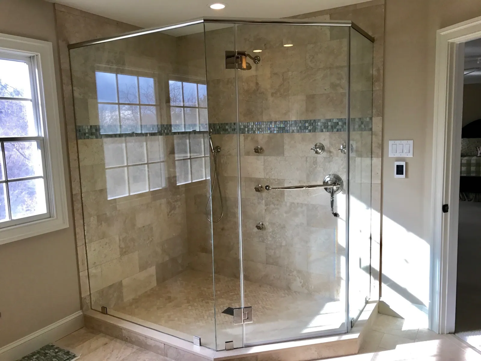 Modern frameless glass shower enclosure with beige tile walls and blue mosaic accent strip in bright bathroom.