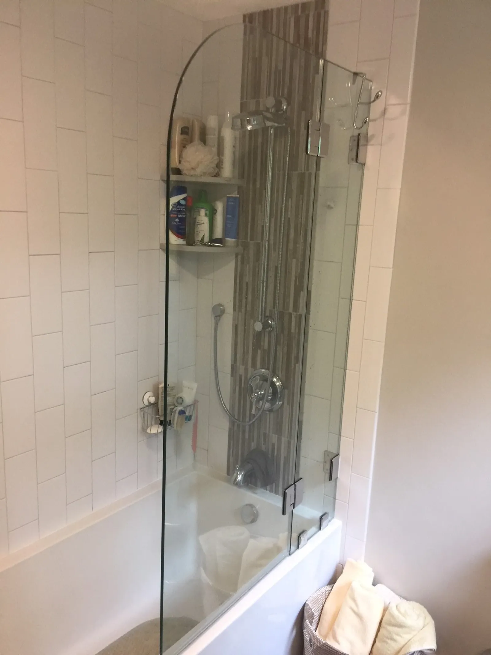 Modern bathroom with glass shower screen, bathtub, tiled wall, showerhead, and toiletries on corner shelves.