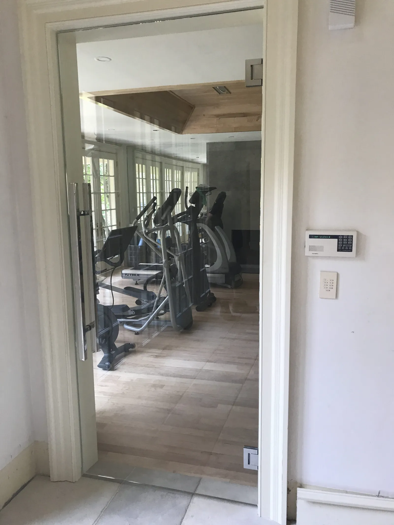 View of a home gym with exercise equipment behind a glass door and security keypad on wall.