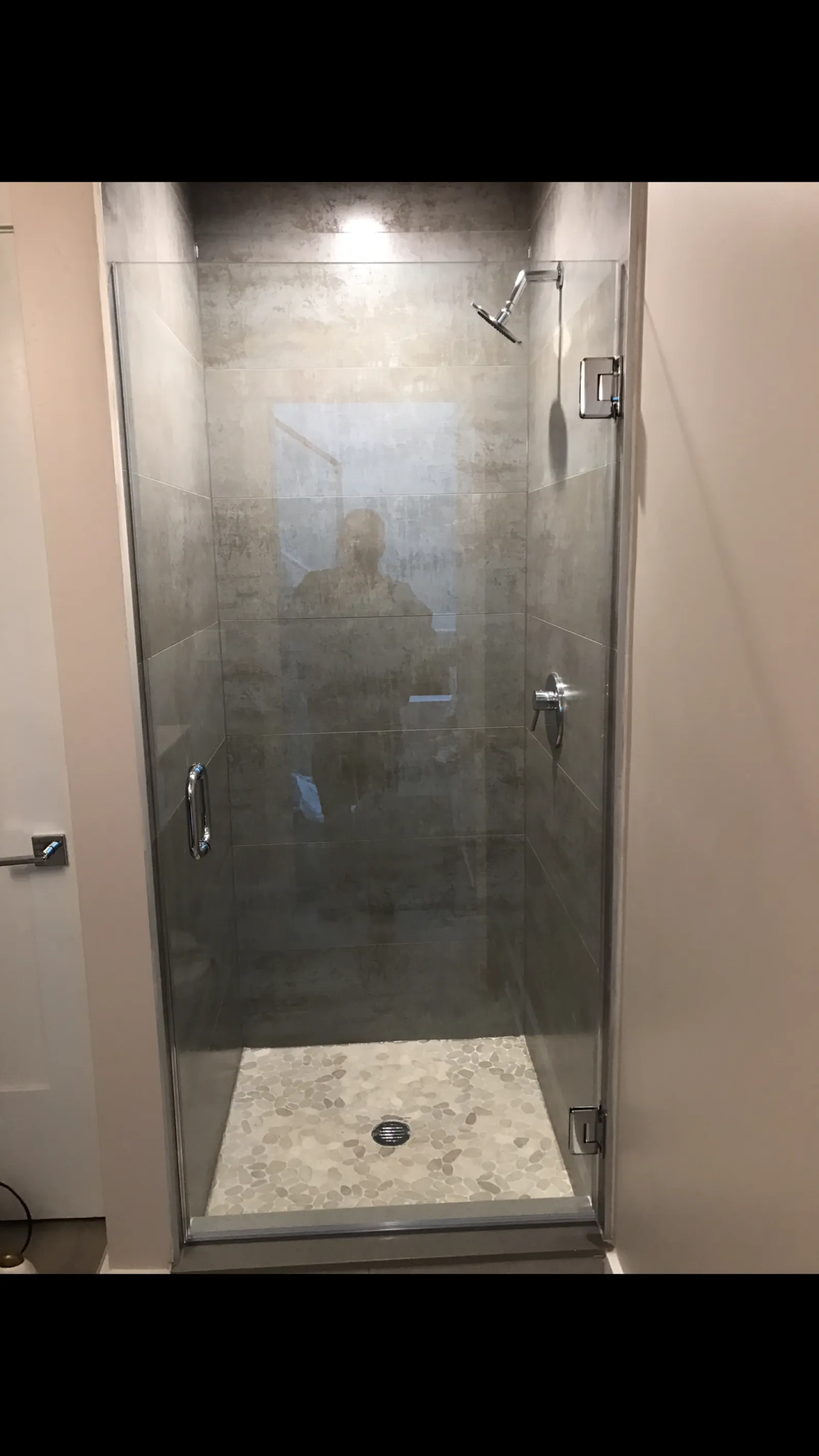 Modern bathroom shower with glass door, gray tiled walls, pebble stone floor, and chrome fixtures.