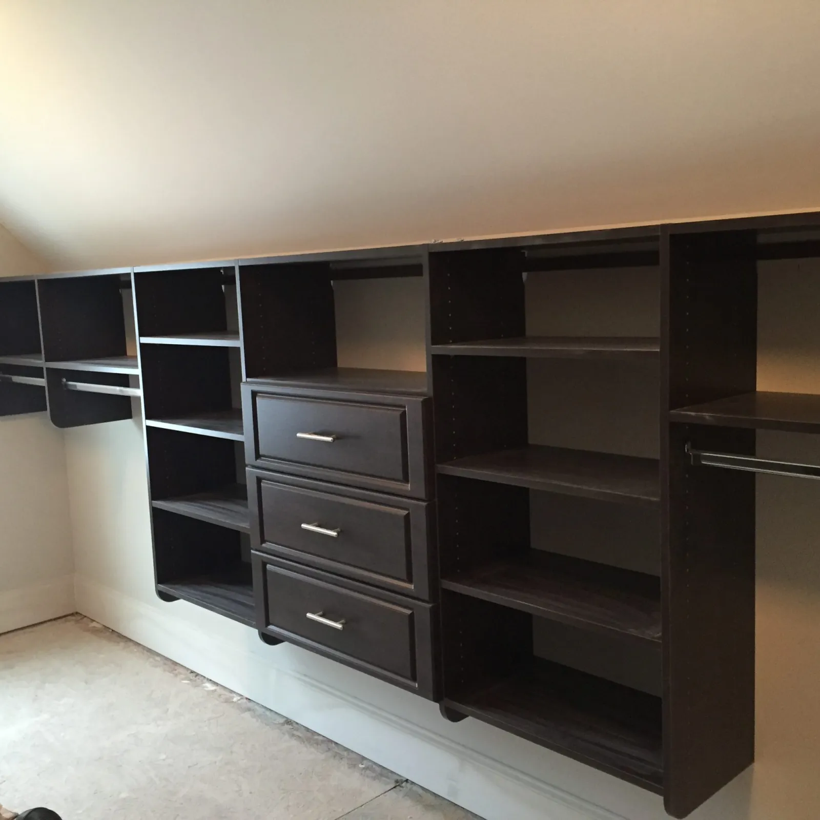 Dark wood floating closet shelves and drawers with metal handles under sloped ceiling in empty room.