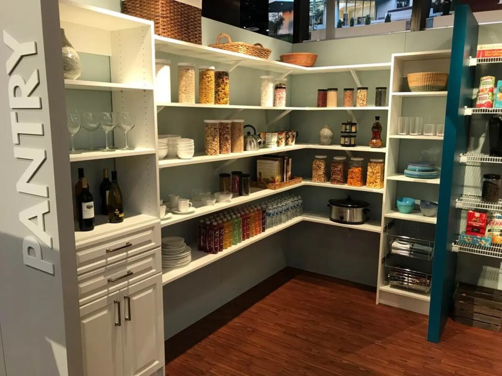 Organized pantry with white shelves holding jars, bottles, dishes, and baskets on wood flooring under bright lighting