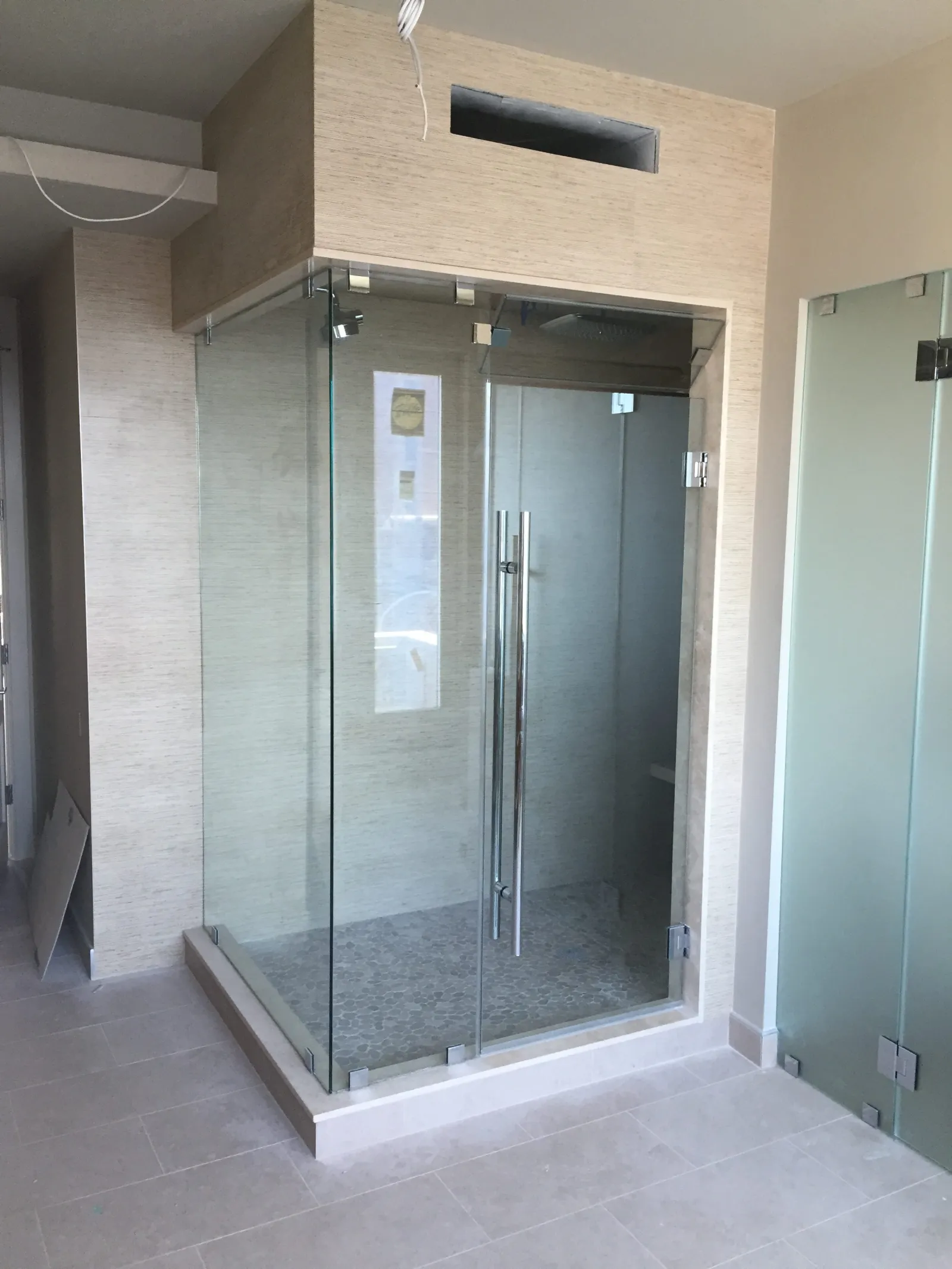 Modern walk-in shower with glass doors in a beige tiled bathroom under construction.