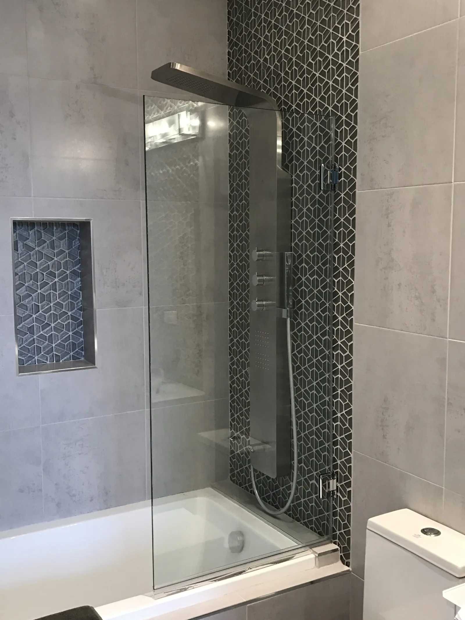 Modern bathroom shower area with glass partition, geometric tile accent, and sleek stainless steel shower panel.