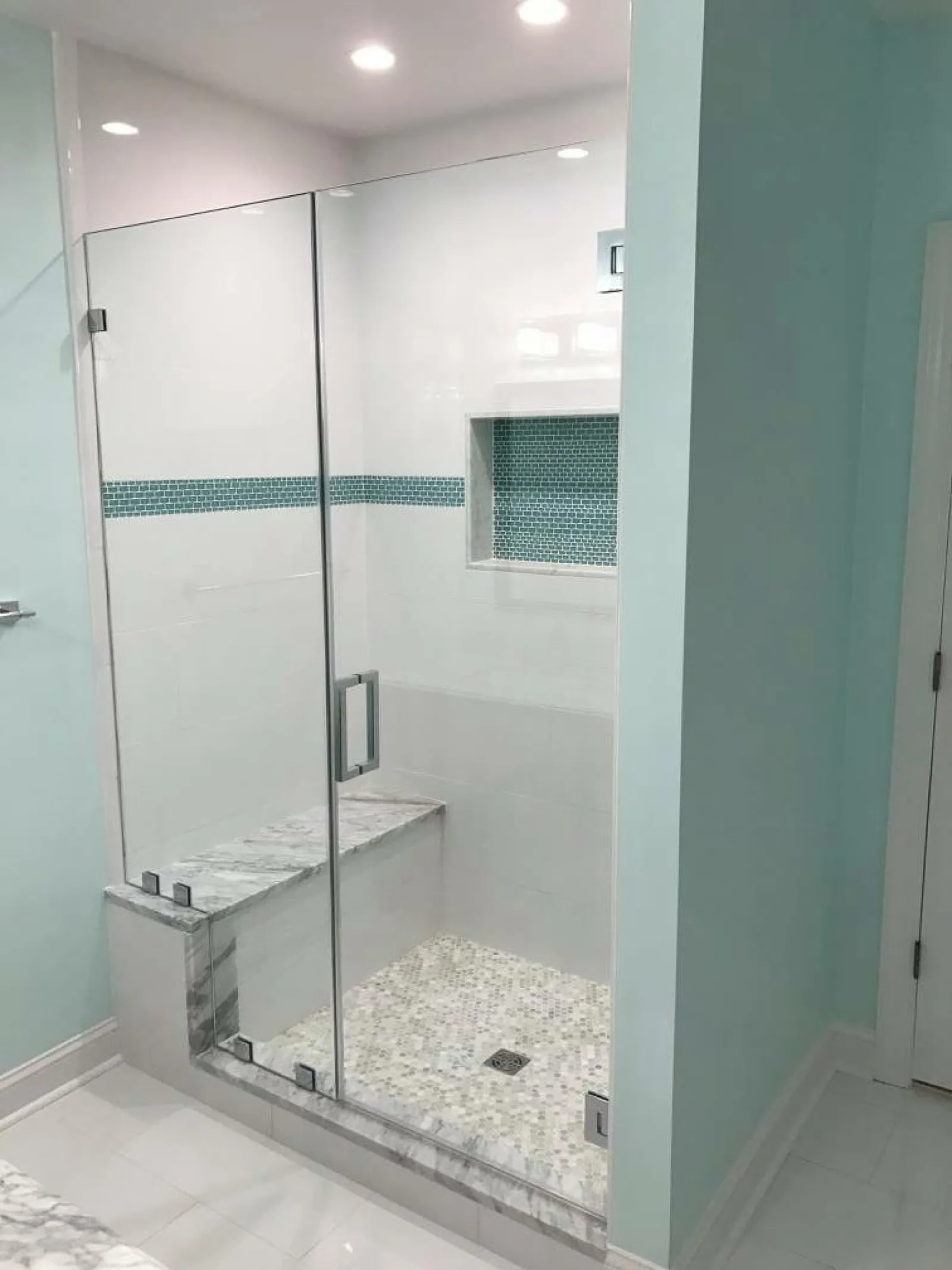 Modern bathroom shower with glass door, built-in bench, mosaic floor tiles, and blue tiled accent strip.