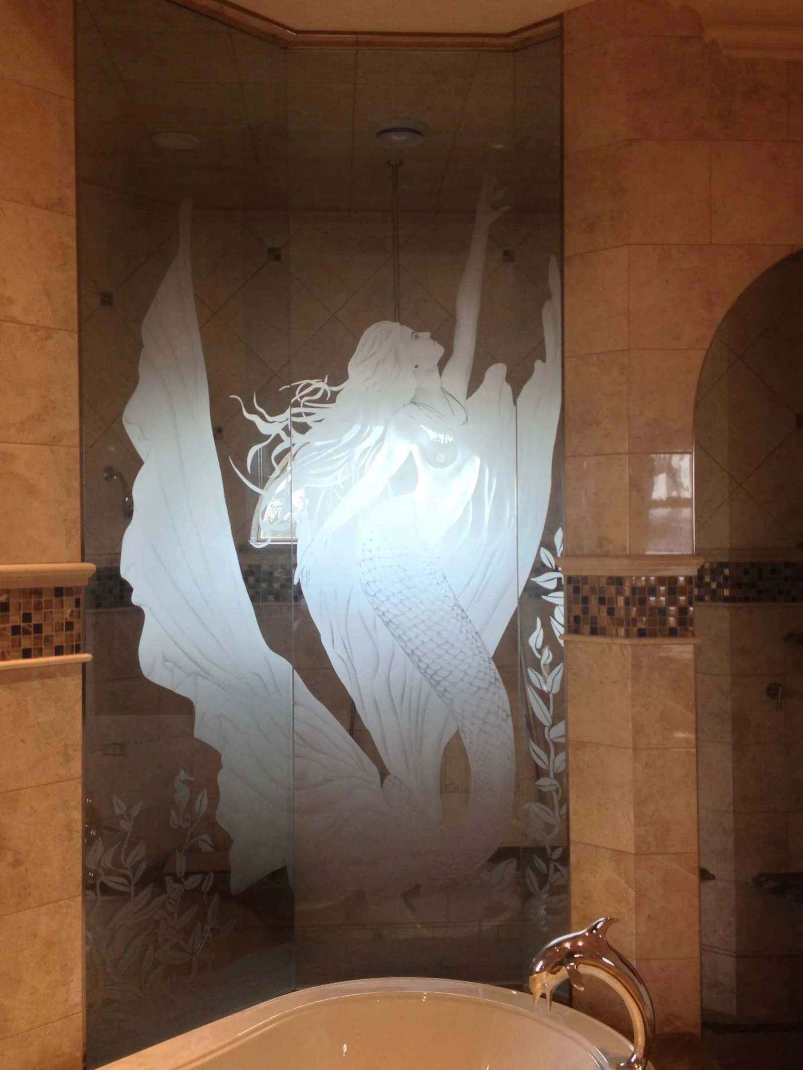Etched glass shower door with a detailed mermaid design in a beige tiled bathroom near a bathtub.