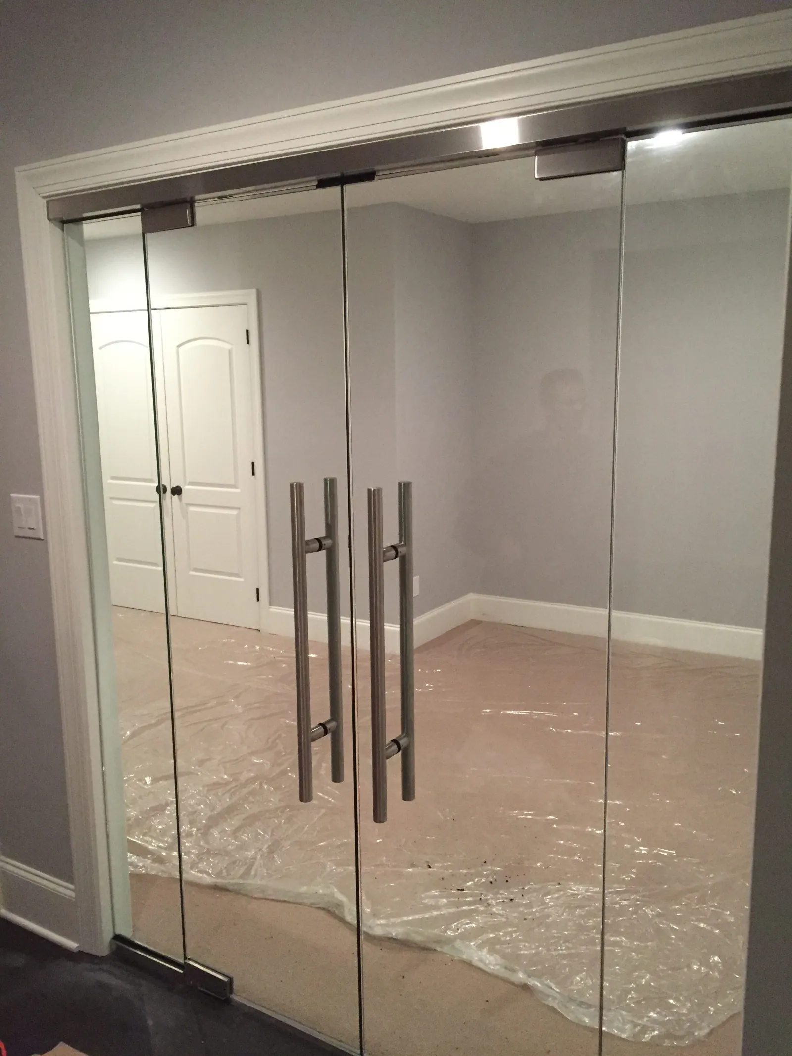 Modern glass double doors with metal handles leading into a room with plastic-covered floor and closed white doors.